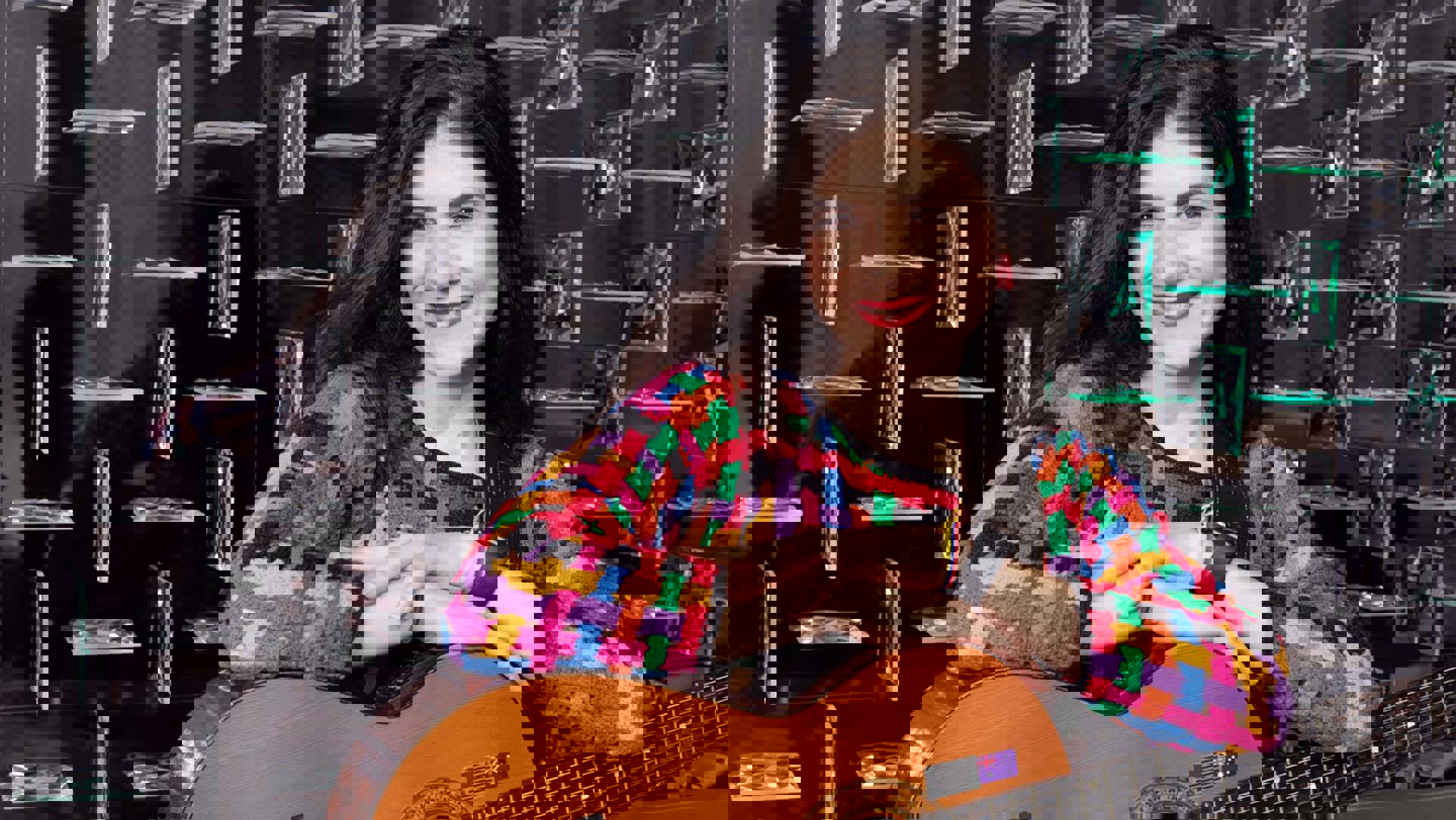 A Colombian woman with long black hair smiles with her arms laid on an acoustic guitar. She is wearing a multi-colored sweater with red lipstick.