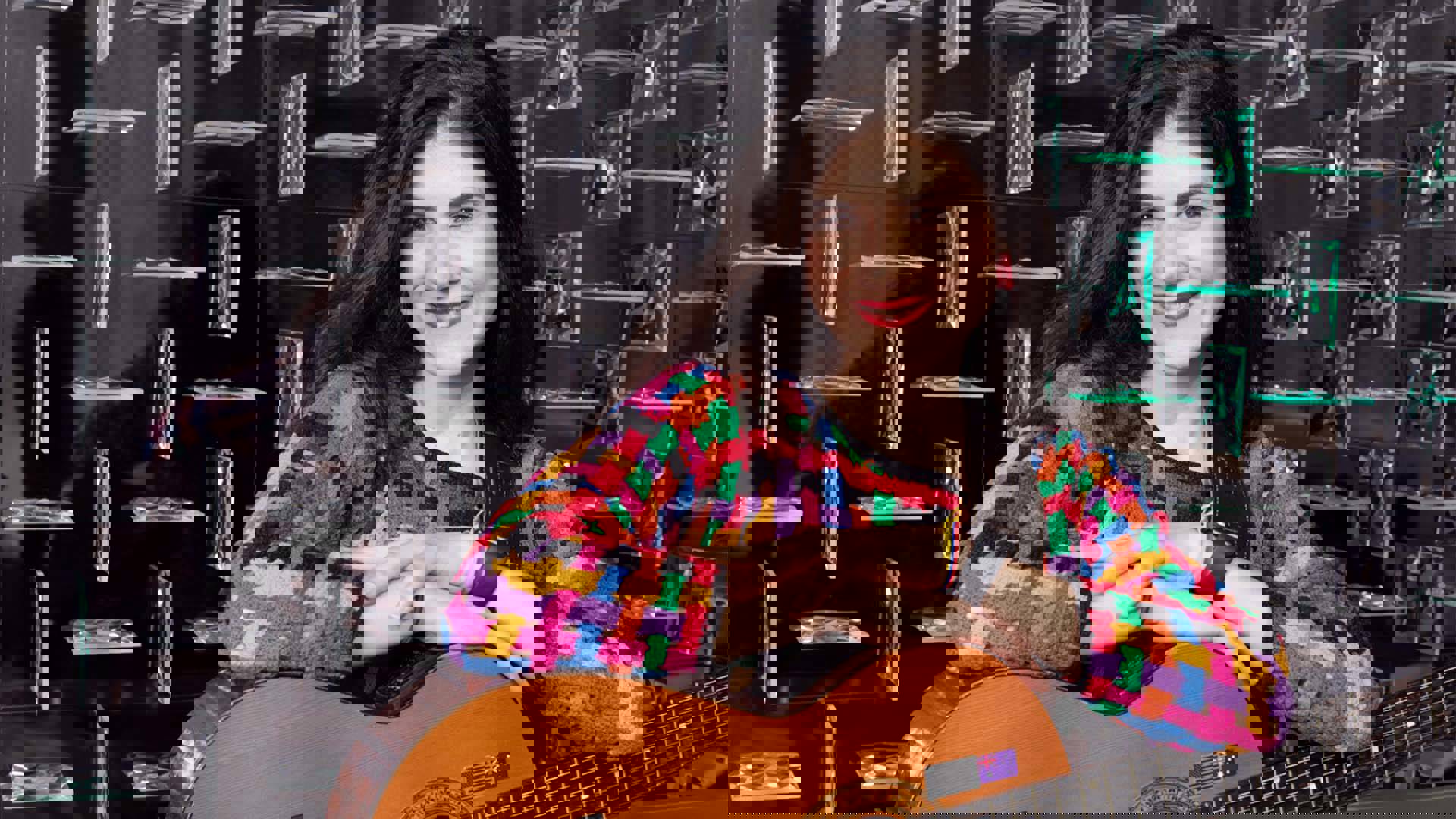 A Colombian woman with long black hair smiles with her arms laid on an acoustic guitar. She is wearing a multi-colored sweater with red lipstick.
