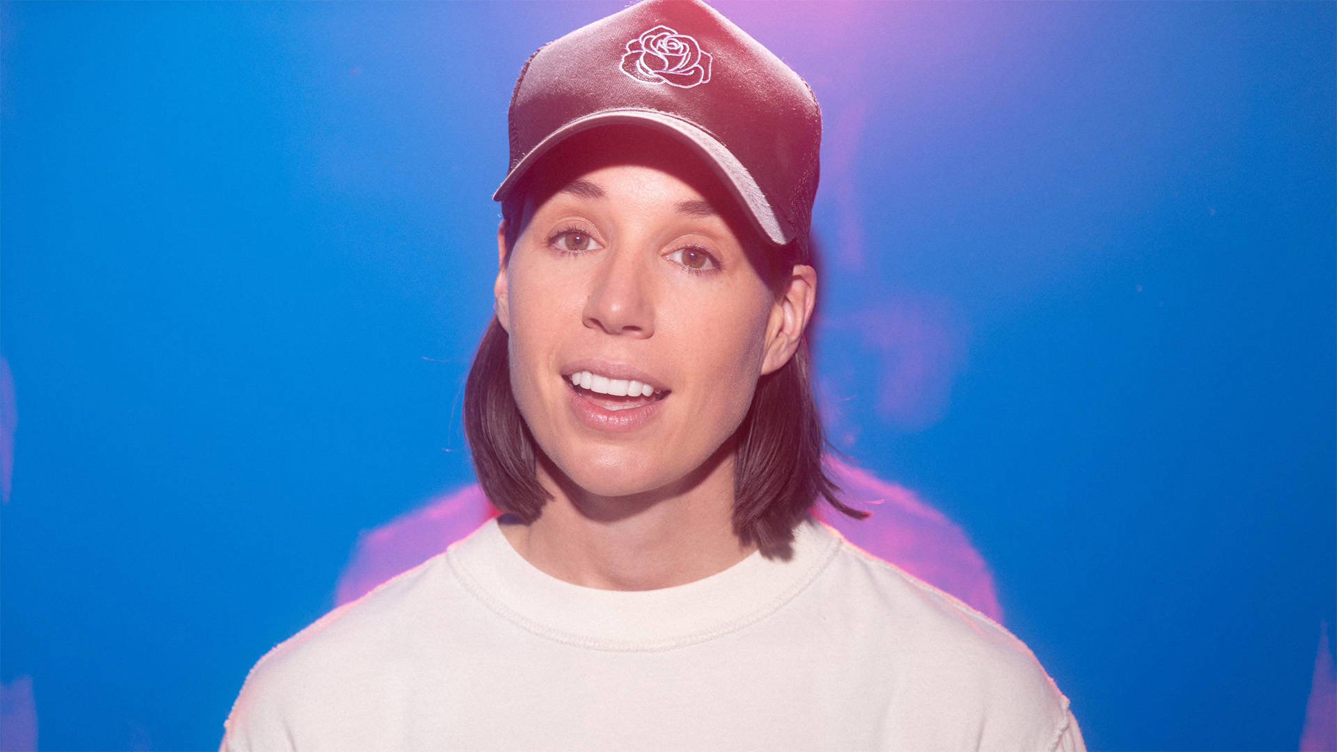 Danae Hays, a woman with light complexion, medium length brown hair, a baseball cap and a white tshirt, slightly smiling towards the viewer with her mouth slightly open.