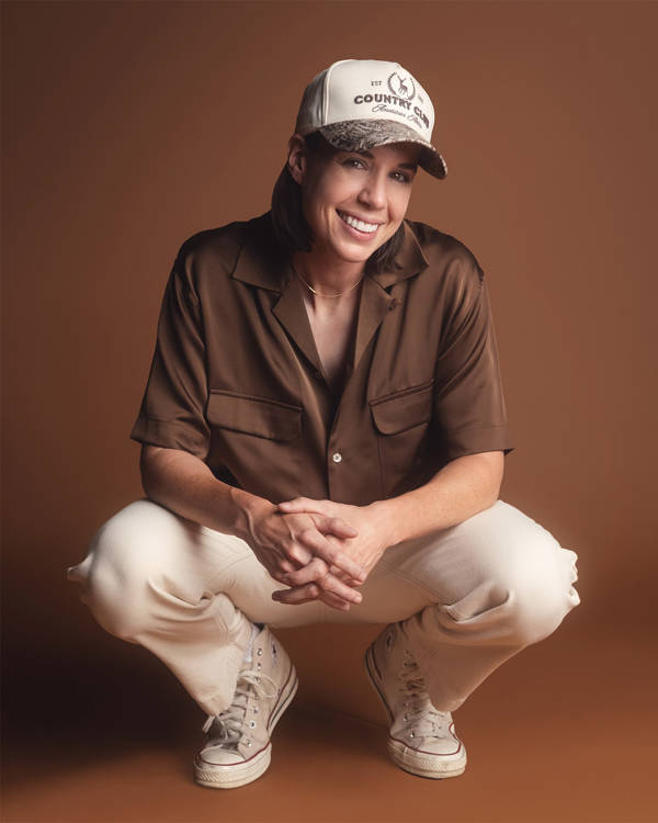 Danae Hays, a woman with light complexion, medium length brown hair, a trucker cap and a baggy brown button up shirt, squatting and smiling towards the viewers