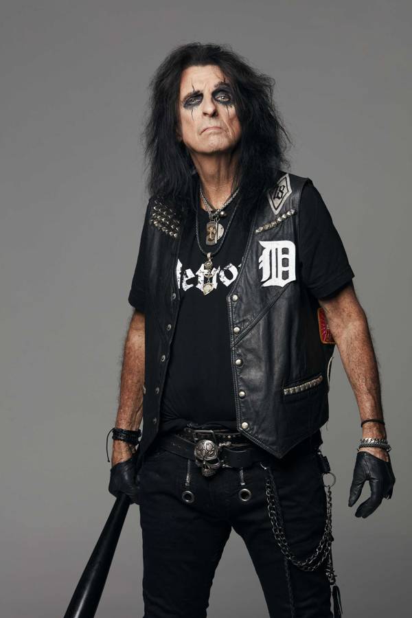 An older man with long, black hair and a lot of eye shadow. He is in black jeans, t-shirt and leather vest and is holding a black baseball bat.