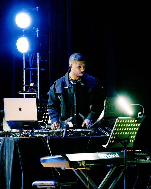 A man with dark complexion, short dark hair and stylish dark clothing DJing on a stage.