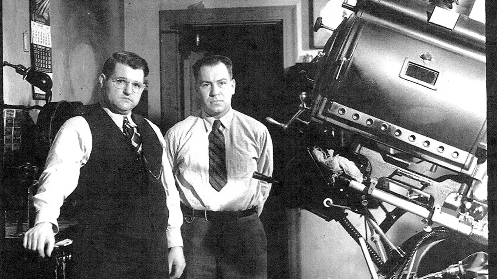 A black and white photo of two men in suits standing next to an old movie projector.