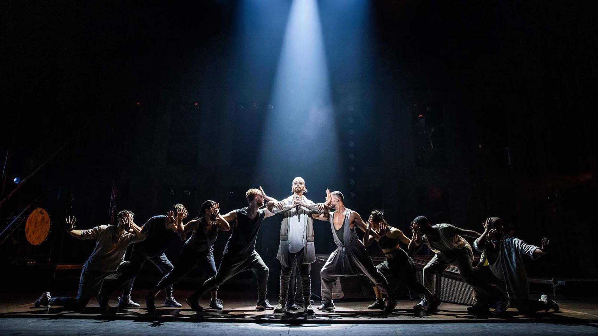 A caucasian man stands in the spotlight with arms linked to cast members on each side in lunge stances.