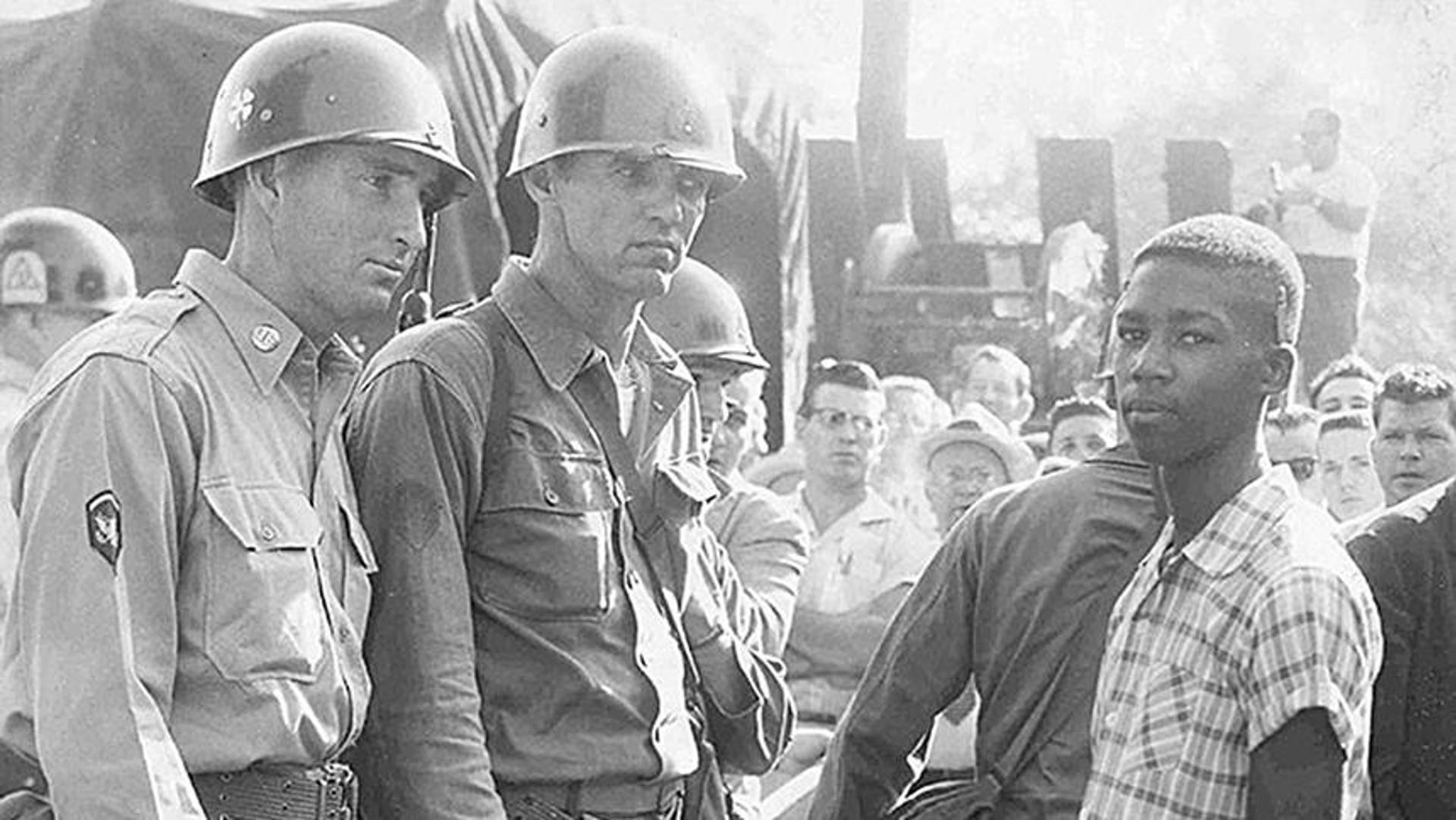 Black and white photo of a young african american man in a plaid shirt standing next to two officers with helmets and uniforms.