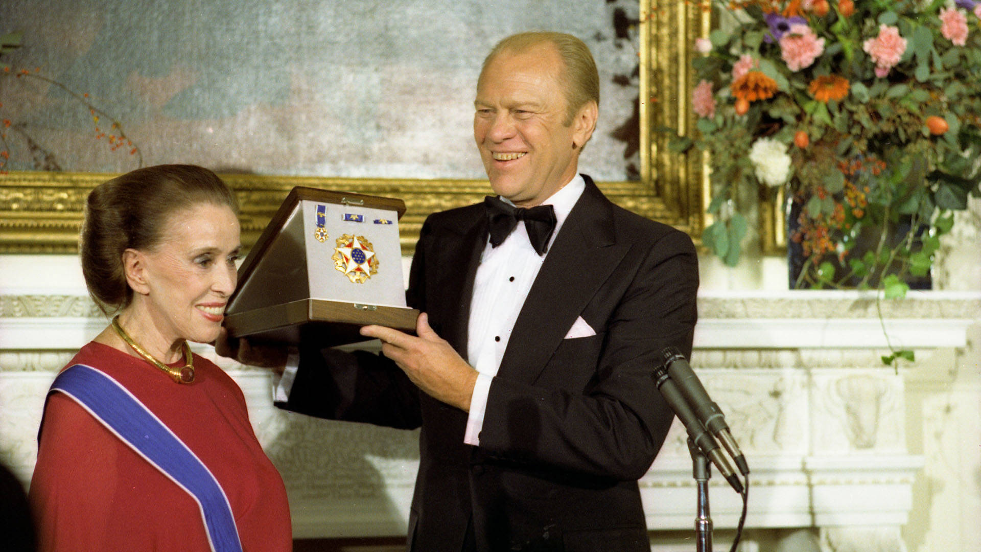 President Gerald Ford, an older caucasian man with short blond hair in a tuxedo presenting a golden medal to Martha Graham, an older caucasian woman with brown hair in a red outfit with a blue sash.