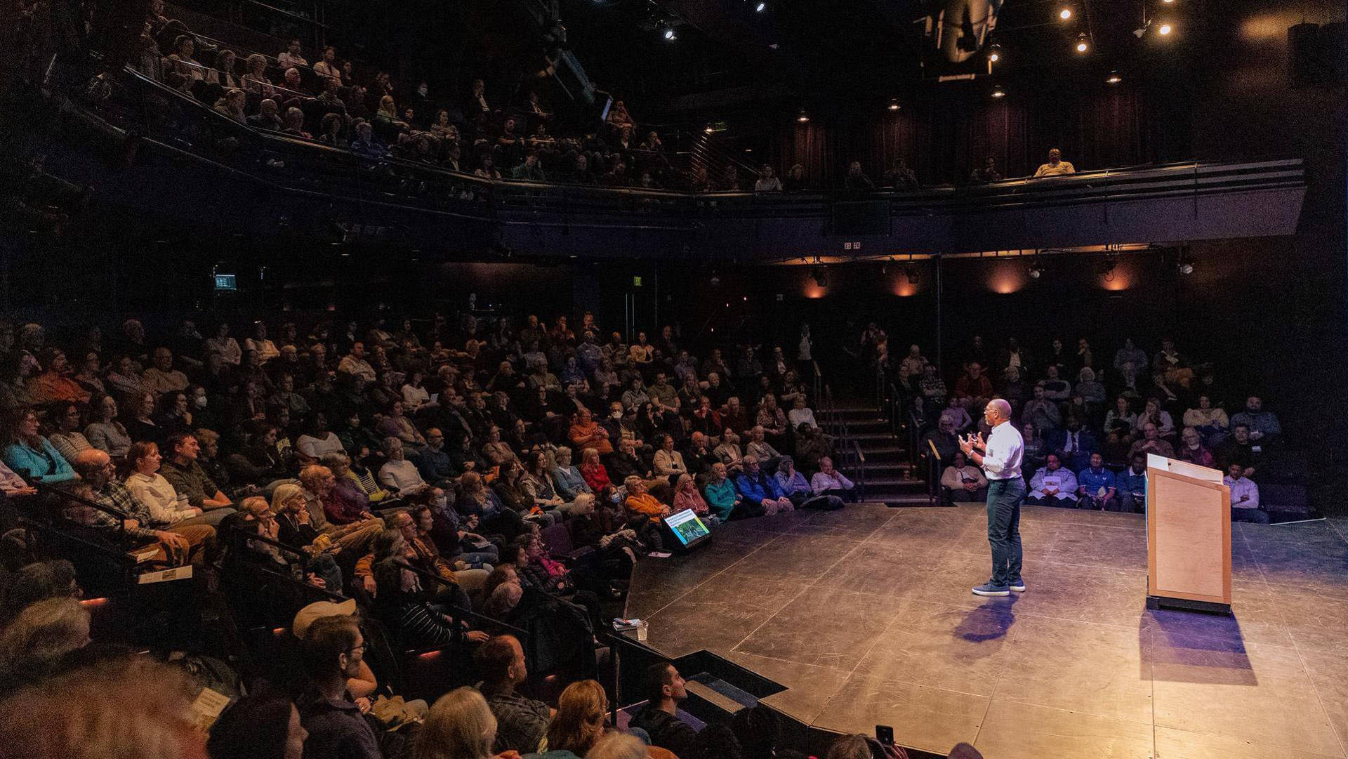 An african american man with buzzed greyish hair, glasses, and a white button-up shirt speaking to a seated audience
