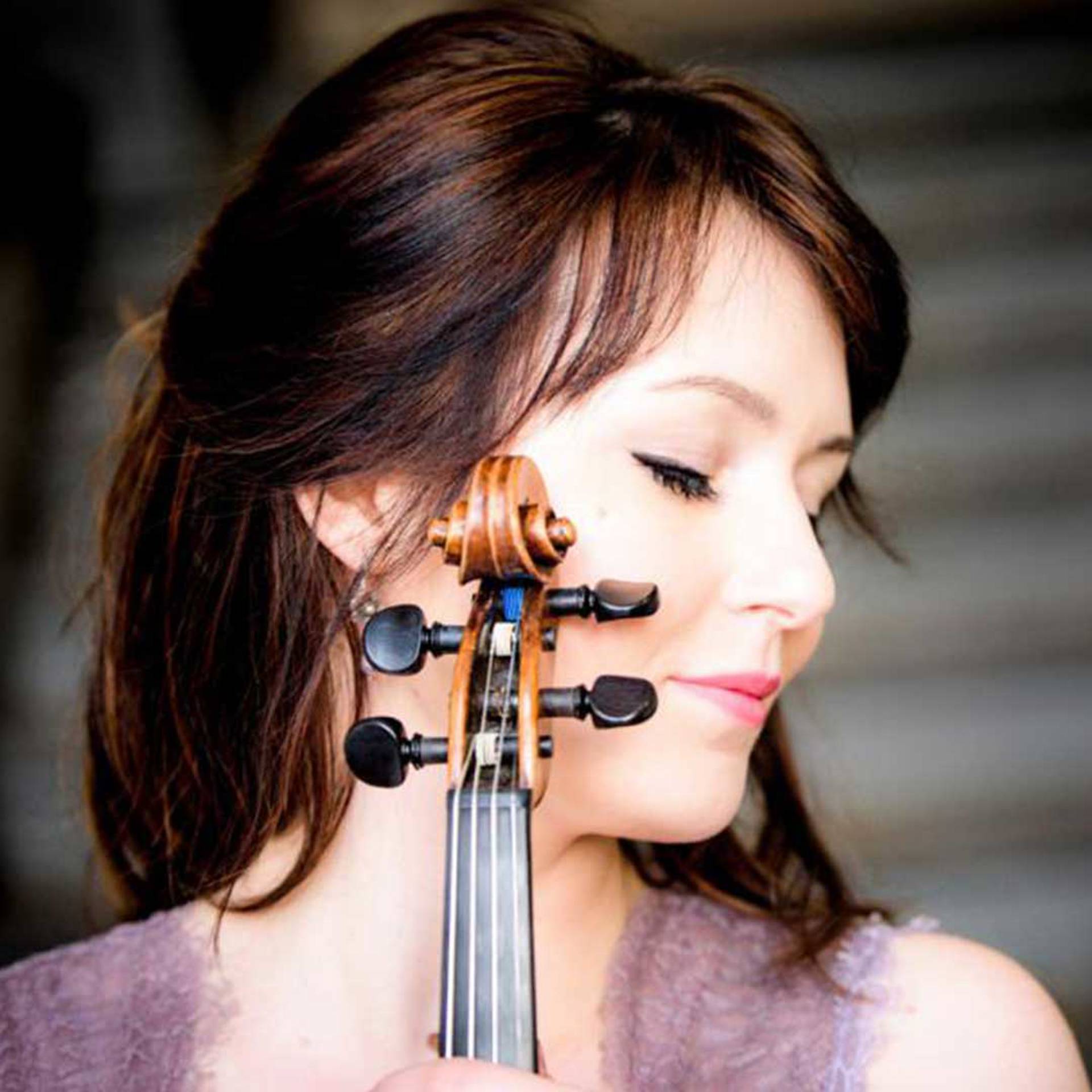 A young adult caucasian woman with dark hair with red highlights turns her head to the side and closes her eyes. Part of a violin is in the frame.