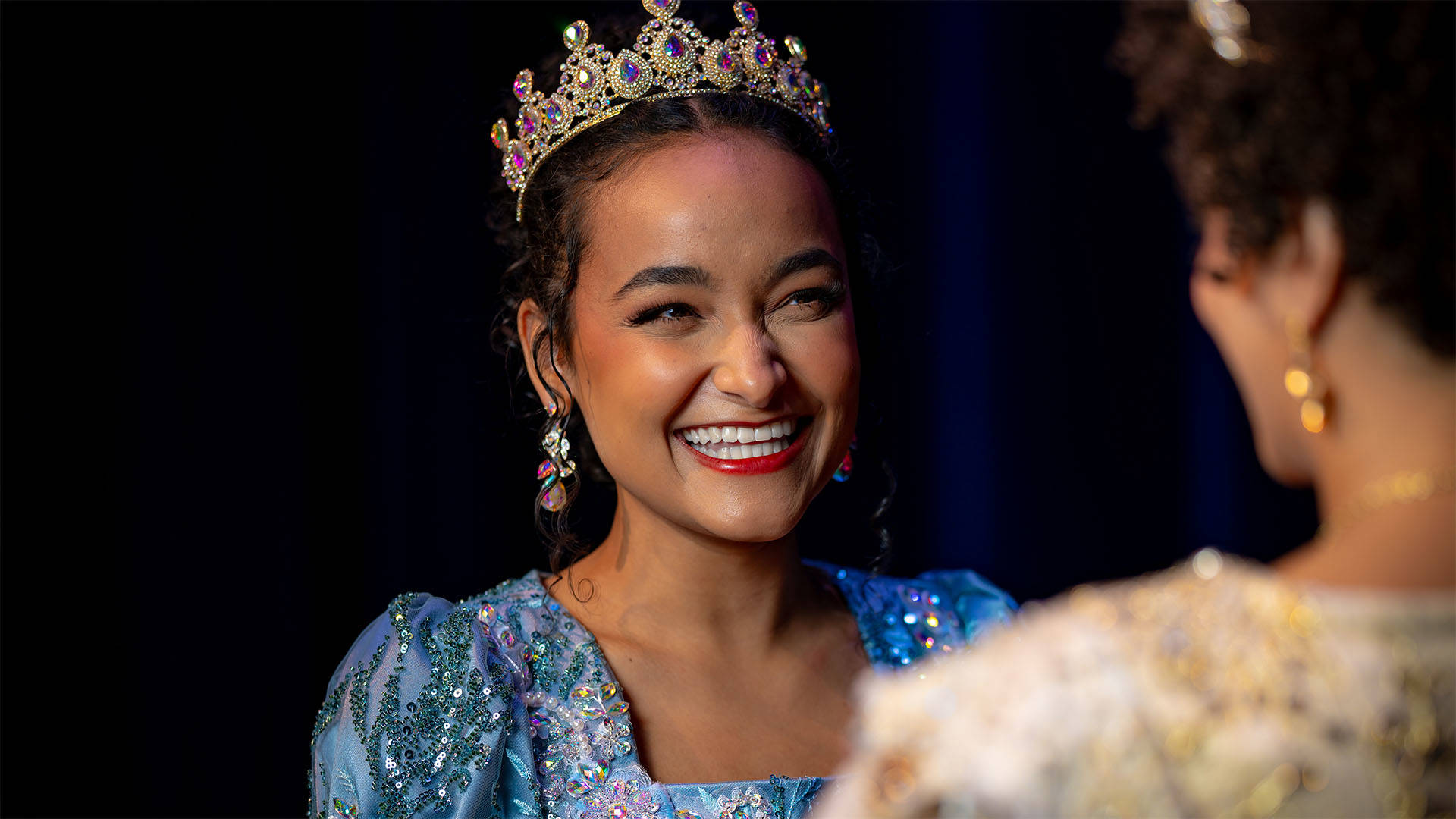 A young woman with medium complexion, dark hair, a sparkling blue dress and a crown smiles towards another woman in the foreground who is out of focus.