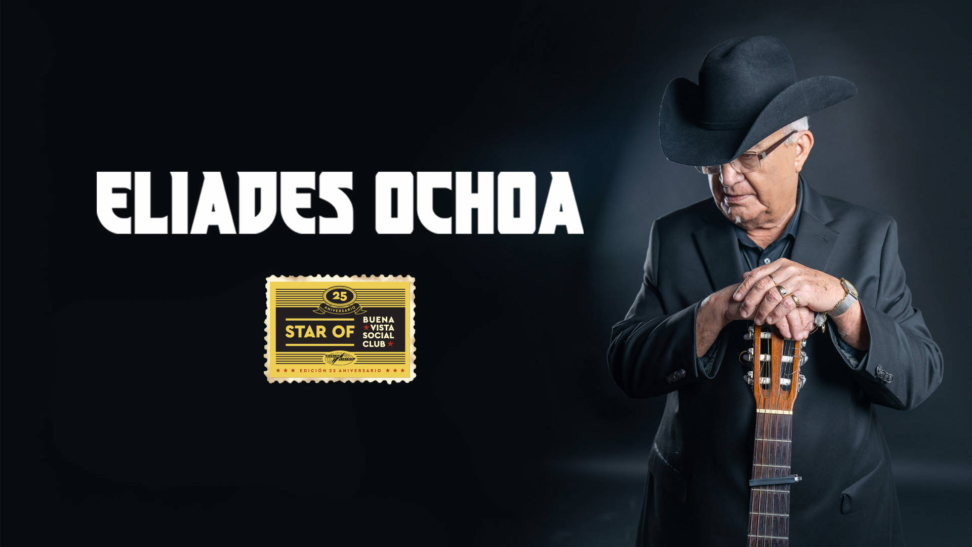 Eliades Ochoa. Star of Buena Vista Social Club. Headshot of Eliades against a black background. An older Cuban man with medium skin tone and short white hair. He is wearing glasses, a black cowboy hat and an all black suit leaning his hands on the top of a guitar.