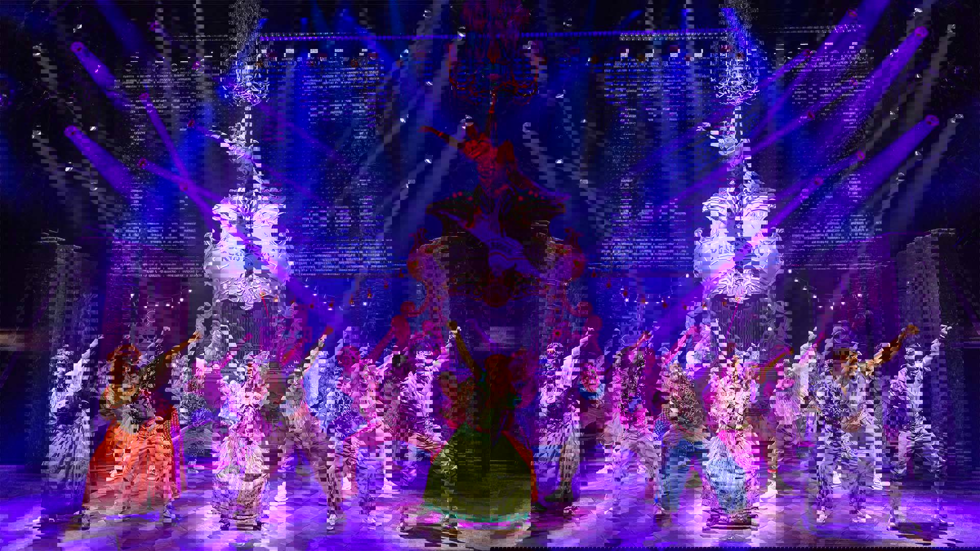 A woman in a pink bomber jacket hangs from a raised chandelier, her left leg and arm both extended as she leans back. Under her, a woman stands on stage, her knees bent and right arm raised upwards as she dances. The stage is filled with dancers in a similar stance, with purple spotlights pointed at them.