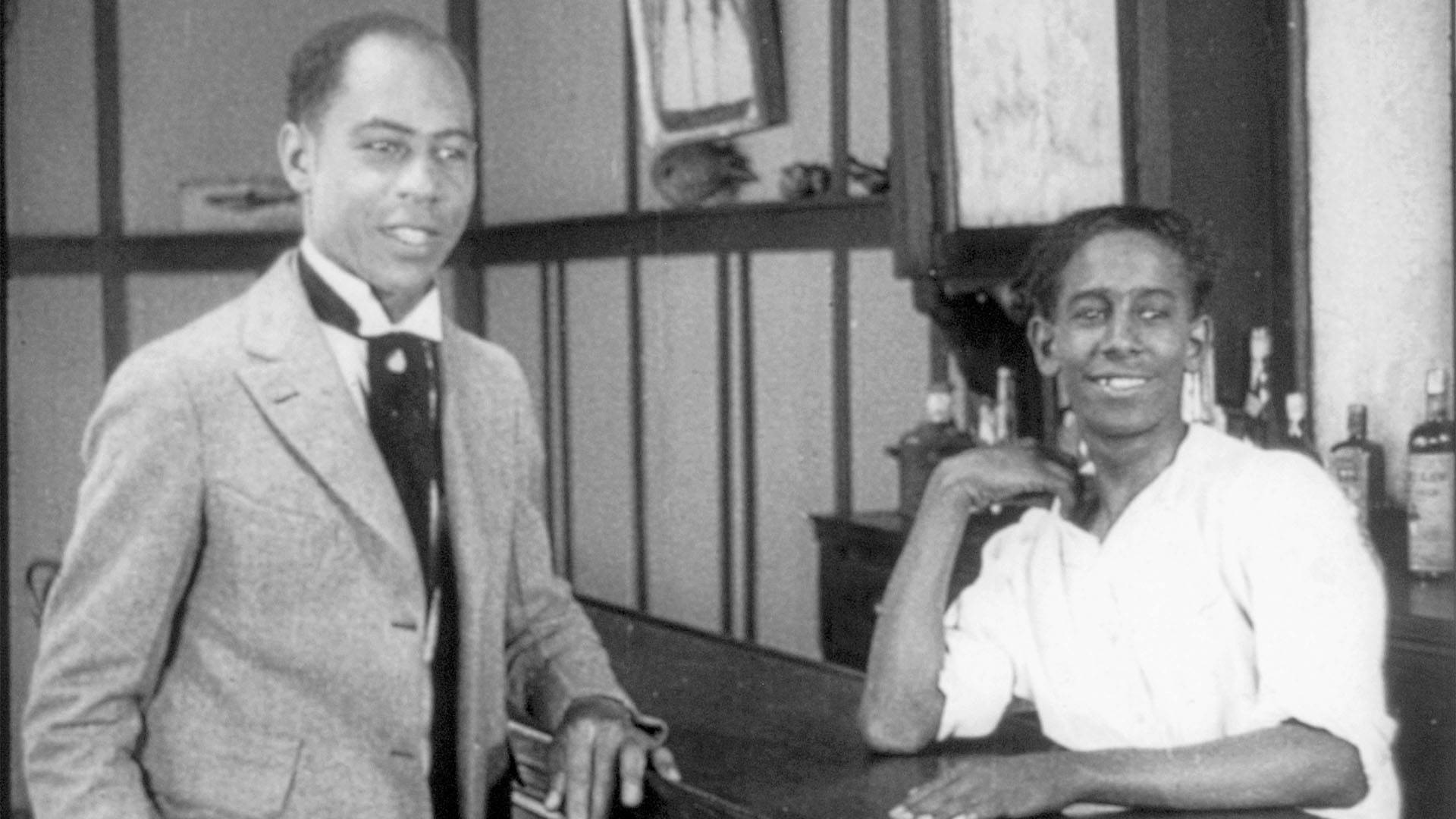 Two Black men in dress clothes leaning against a bar, smiling. A black and white still from "Ten Nights in a Bar Room."
