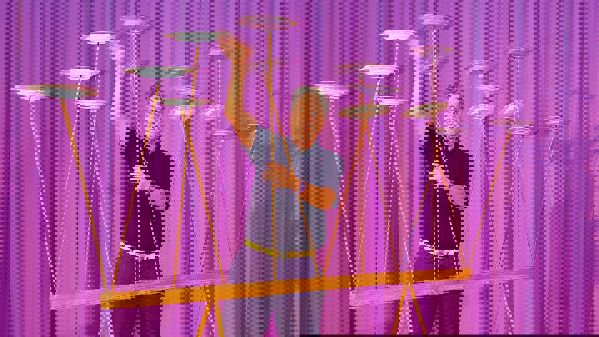 A man dressed in all purple with short grey hair keeping 13 plates spinning on wooden stilts.