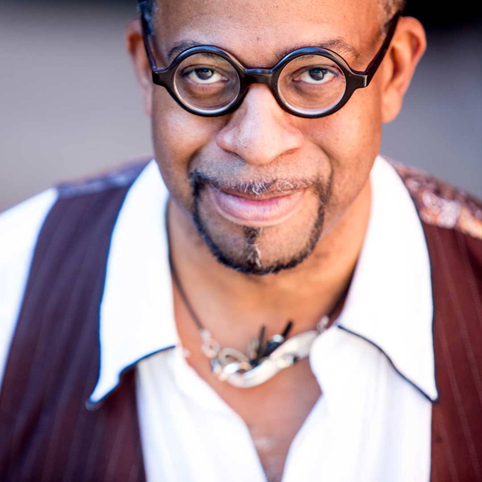 Headshot of a middleaged african american man with a goatee and glasses. He is wearing a collared white shirt with a burgundy vest as well as a necklace.