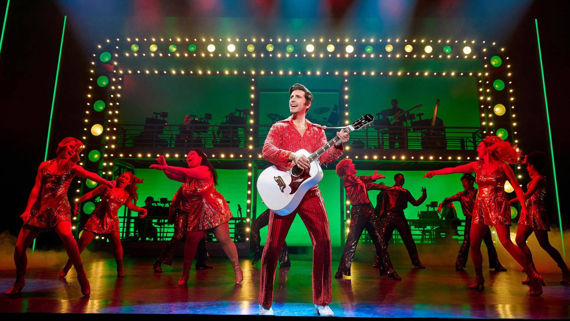 A man with light complexion, greased brown hair and a red outfit performing with an acoustic guitar. Dancers in red light are behind him.