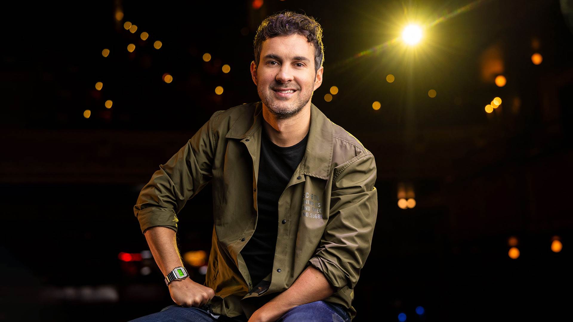 A young caucasian man with short brown hair in a black shirt and olive green button up shirt over sitting and smiling to the camera. Stage lights shine behind him.