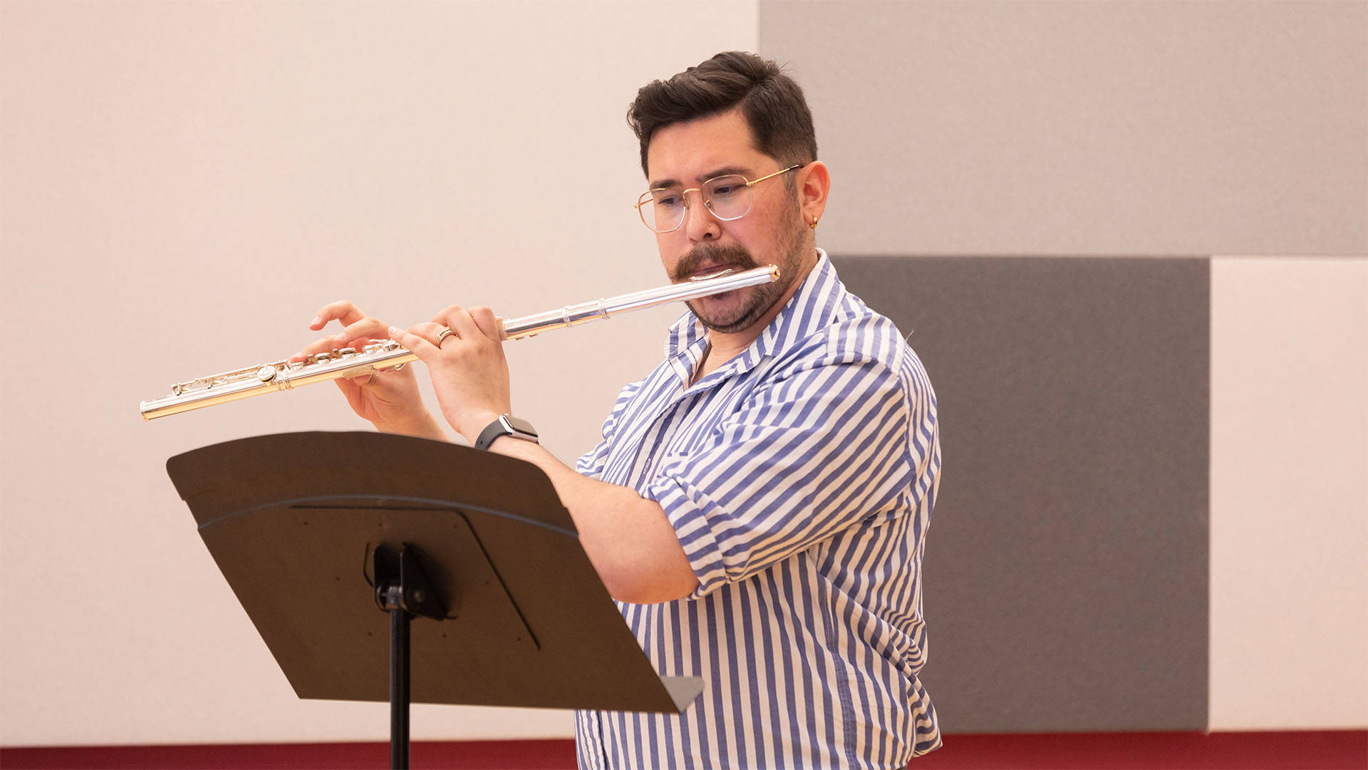 A man with medium complexion, short brown hair, and facial hair in a striped button up shirt playing flute.