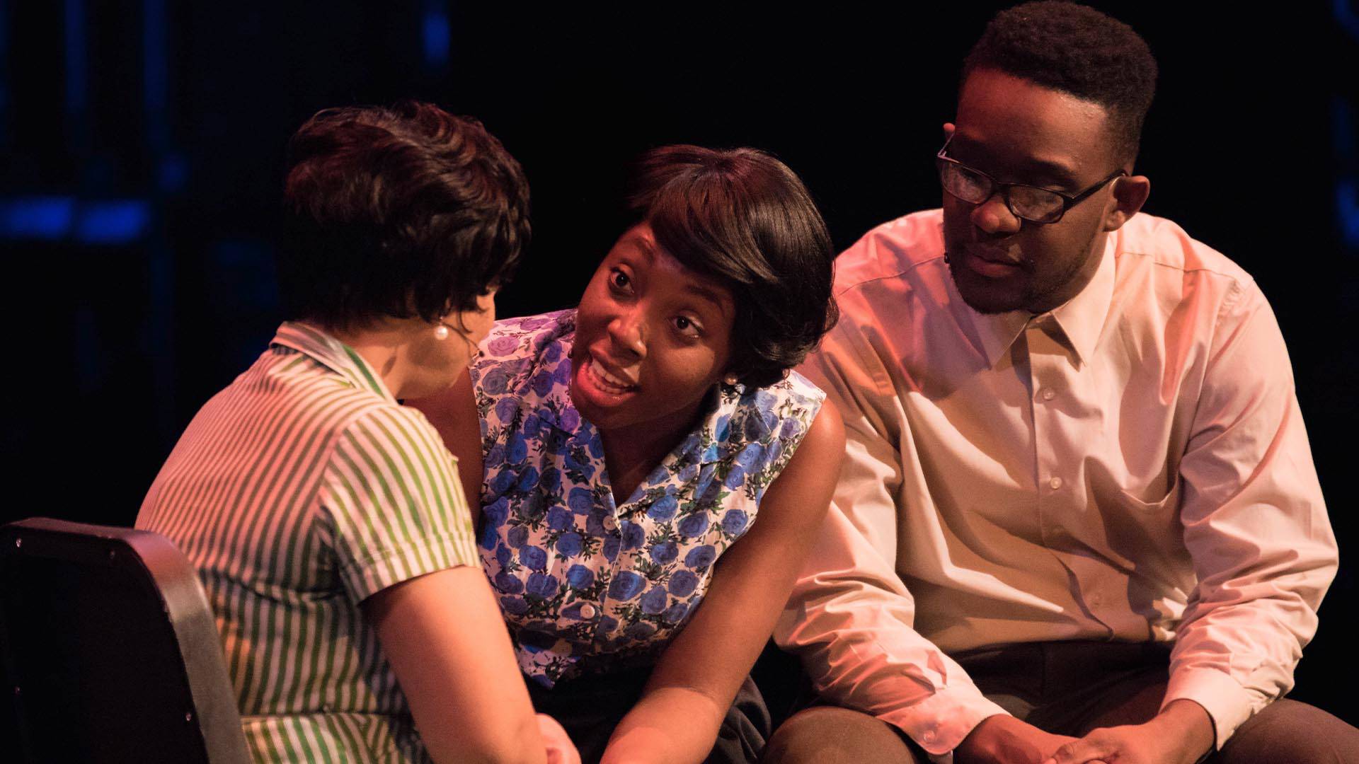 Two Black women in period dresses having a conversation. A Black man sits next to them listening.