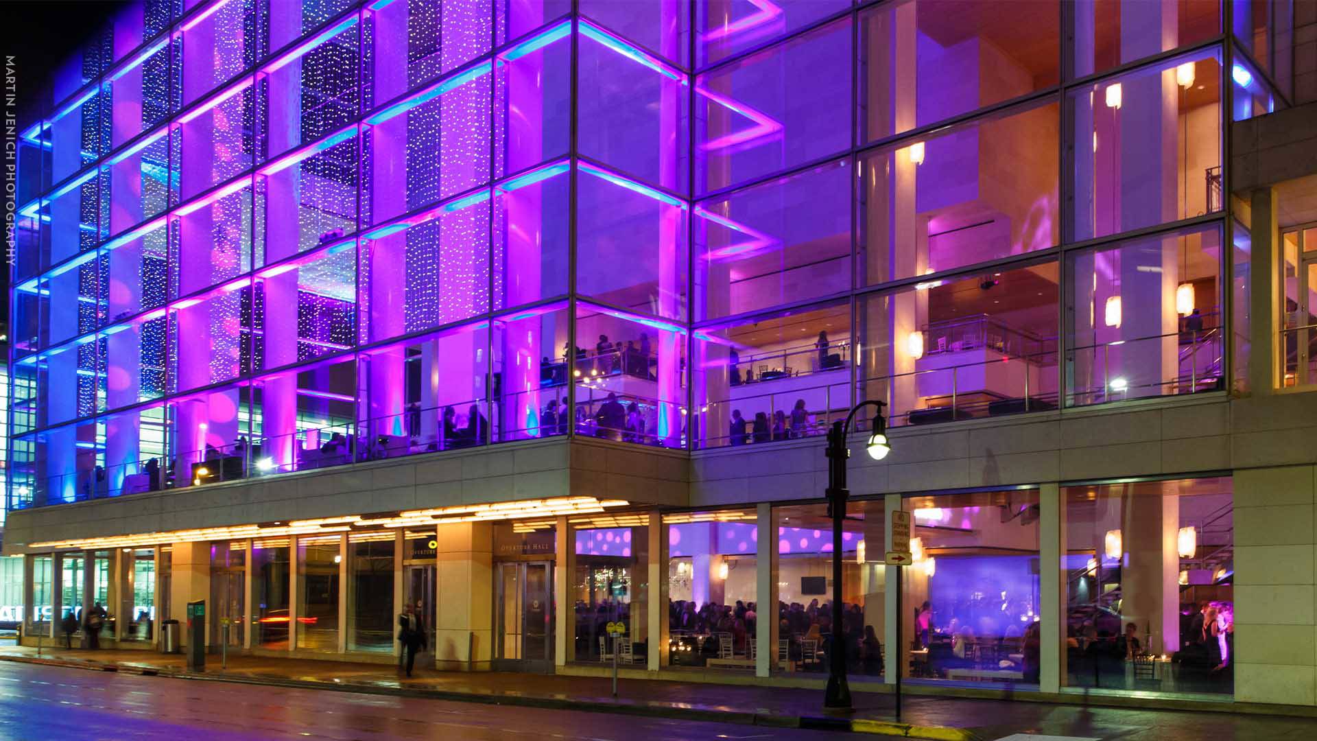The outside of Overture Center at night. The building is like up in purple colors and many people are gathered inside.