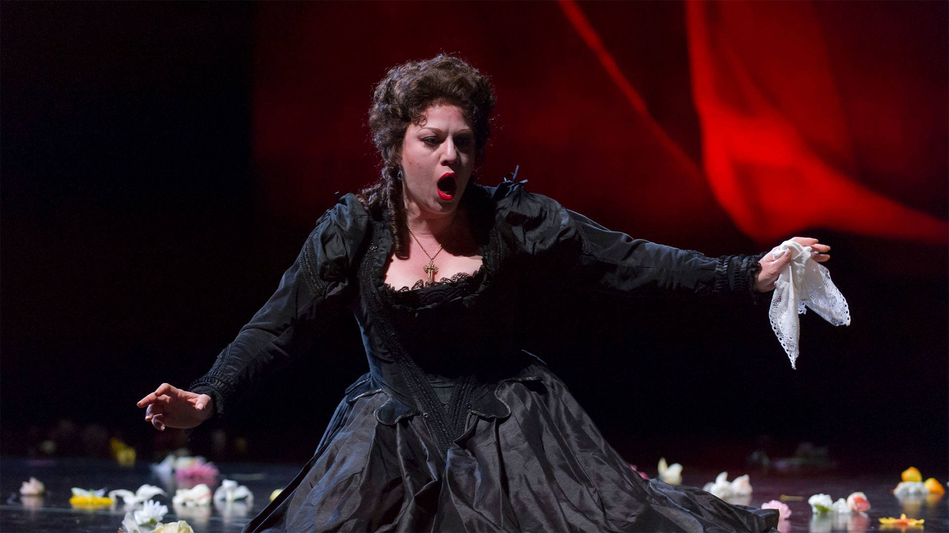 A woman performing in the opera "Don Giovanni". She has a dark dress on, is holding a handkerchief and singing with her arms outstretched.