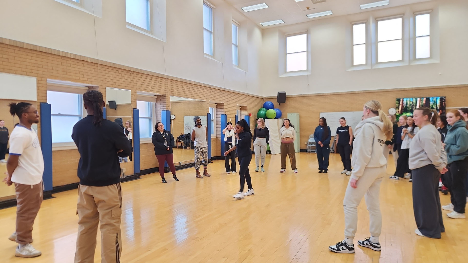 A group of high school students learn dance moves in a bright dance studio with a wood floor.