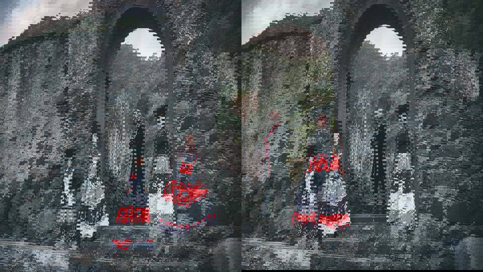 Four people with light complexion stand for a photo in a stone setting with an archway. They are wearing ornate traditional black white and red Ukrainian outfits. Three have large plush black headwear on