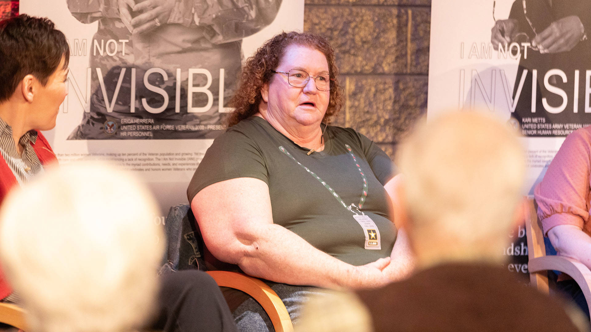 An older woman with light complexion, curly brown hair and a green top sitting and in the act of speaking. Other blurry heads are slightly visible.