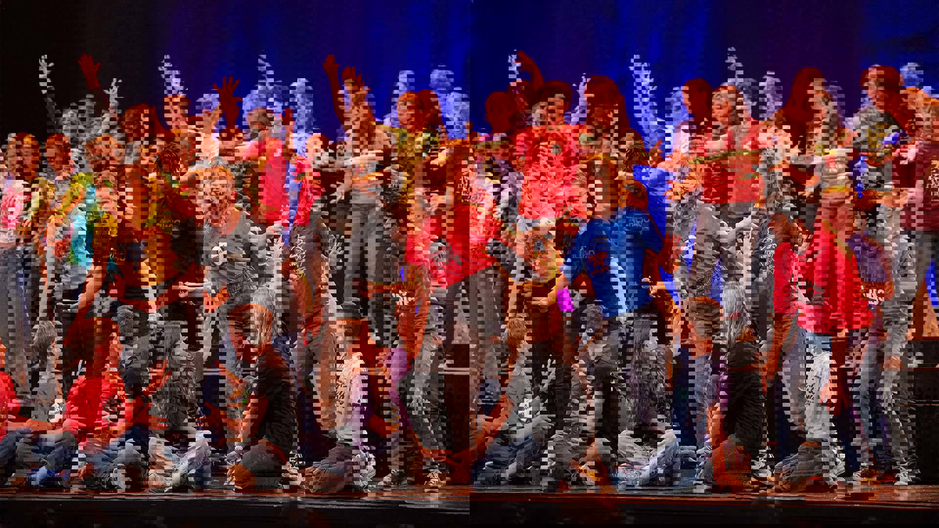 A group of high schoolers in various colorful outfits performing on a dark stage.