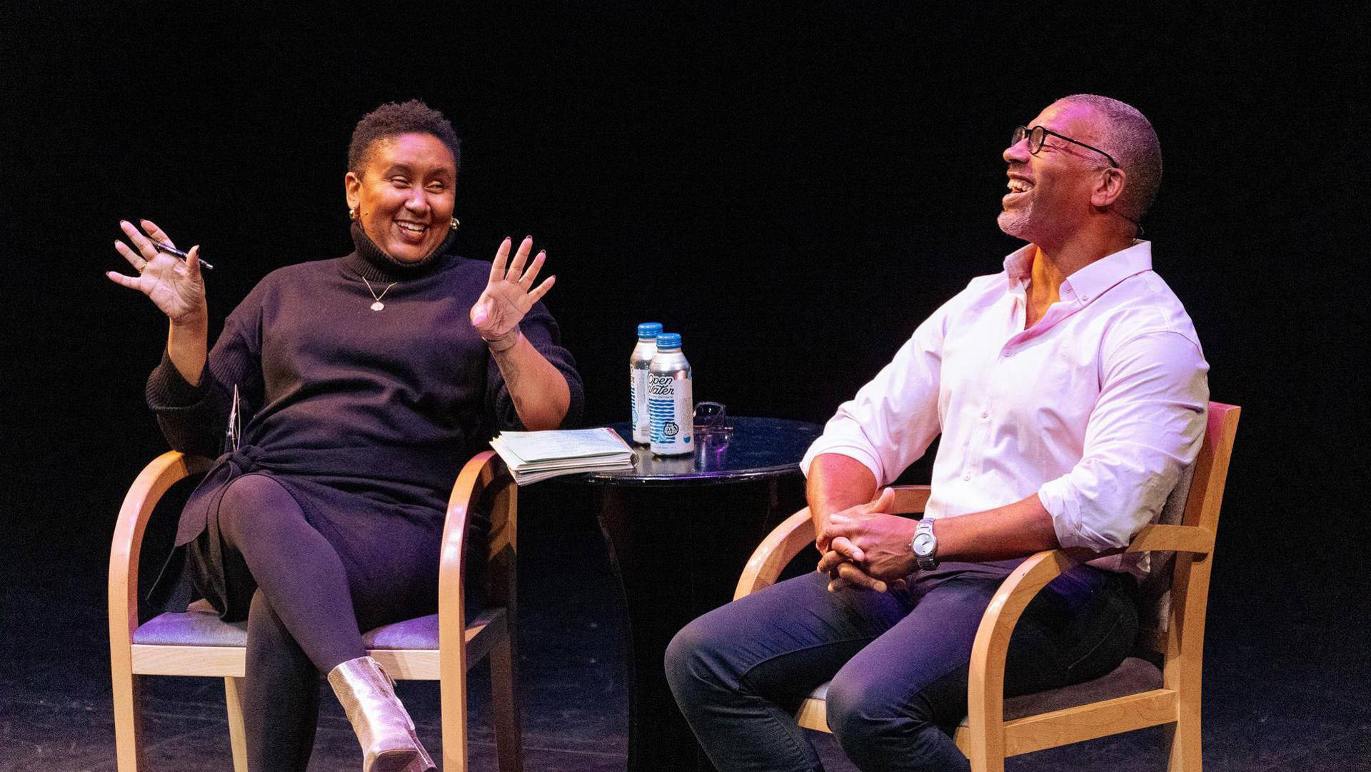 An african american man with buzzed greyish hair, glasses, and a white button-up shirt sitting next to an african american woman with short dark hair and a dark outfit. They are both laughing.