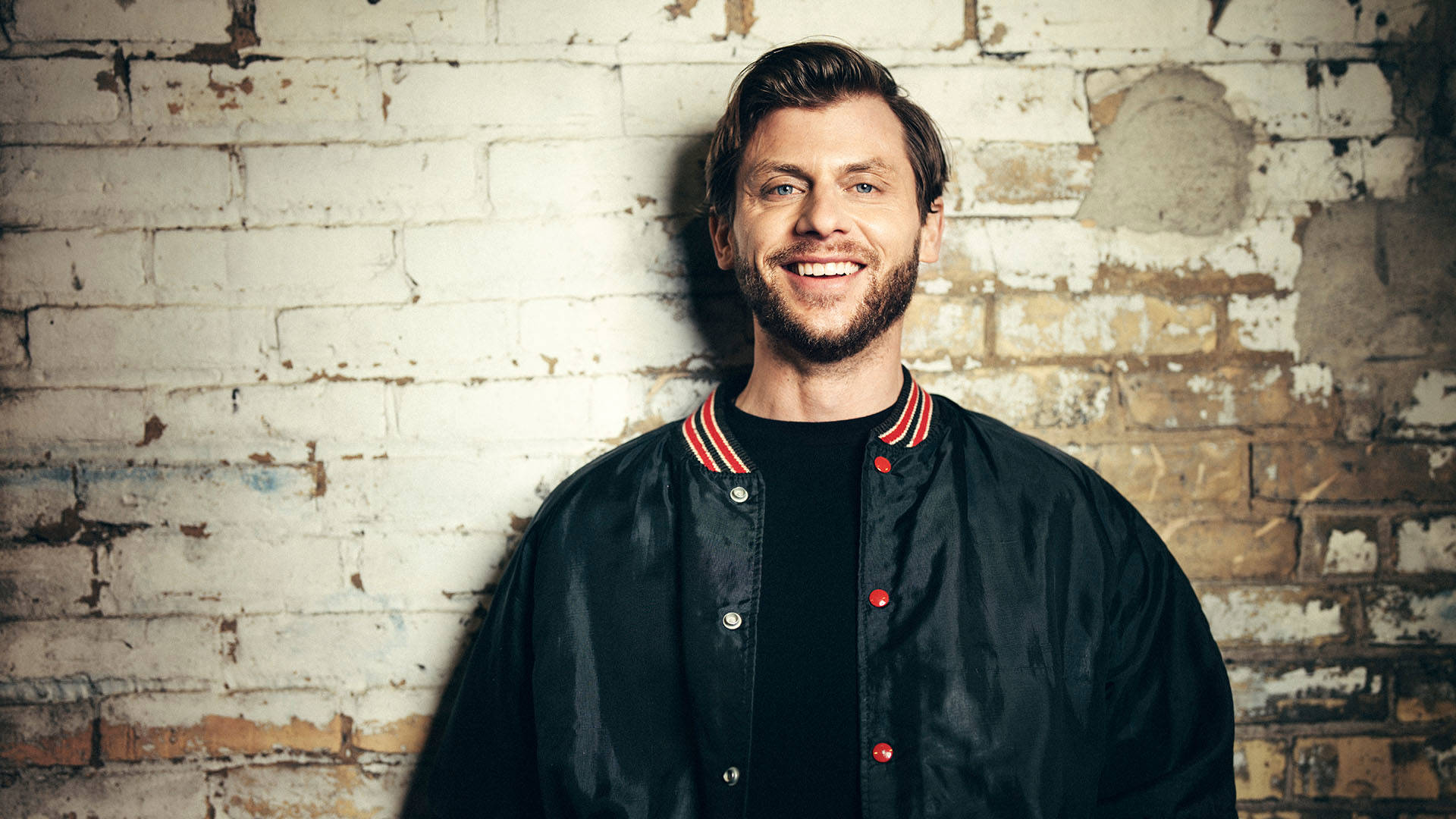 Charlie Berens, a man with light complexion wearing a dark t-shirt and jacket, smiling at the camera in front of a whitewashed brick wall.