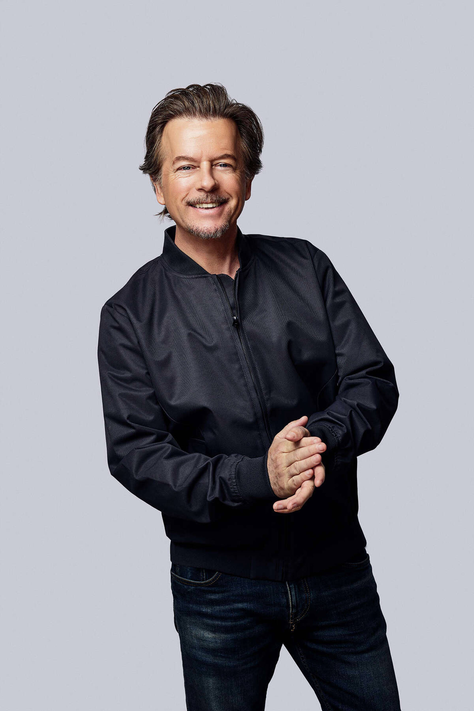 David Spade headshot. A caucasian man smiling with grayish blonde hair and facial hair wearing a black bomber jacket and jeans. His hands are clasped together in front.