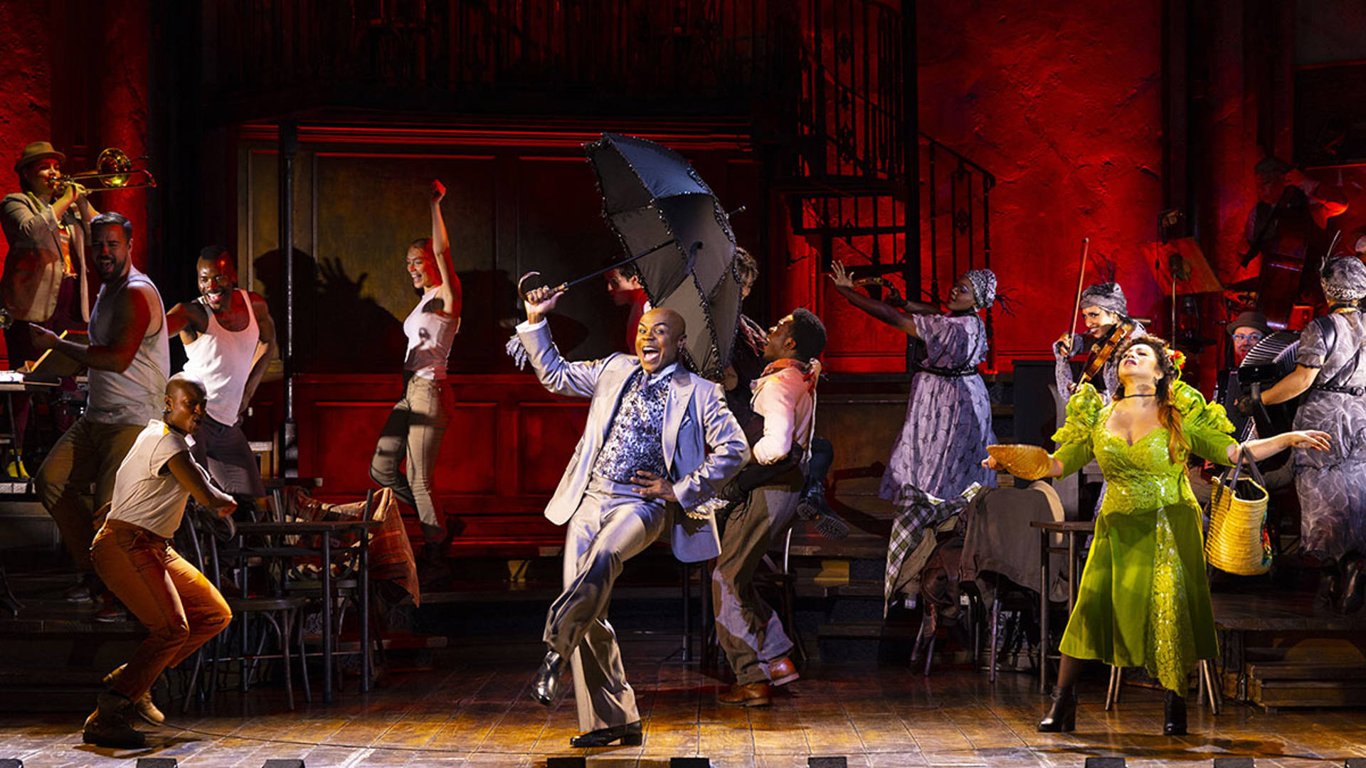 The cast of Hadestown performing on stage. One man in a silver suit holds an open umbrella up. A woman in a lime green dress is dancing nearby.