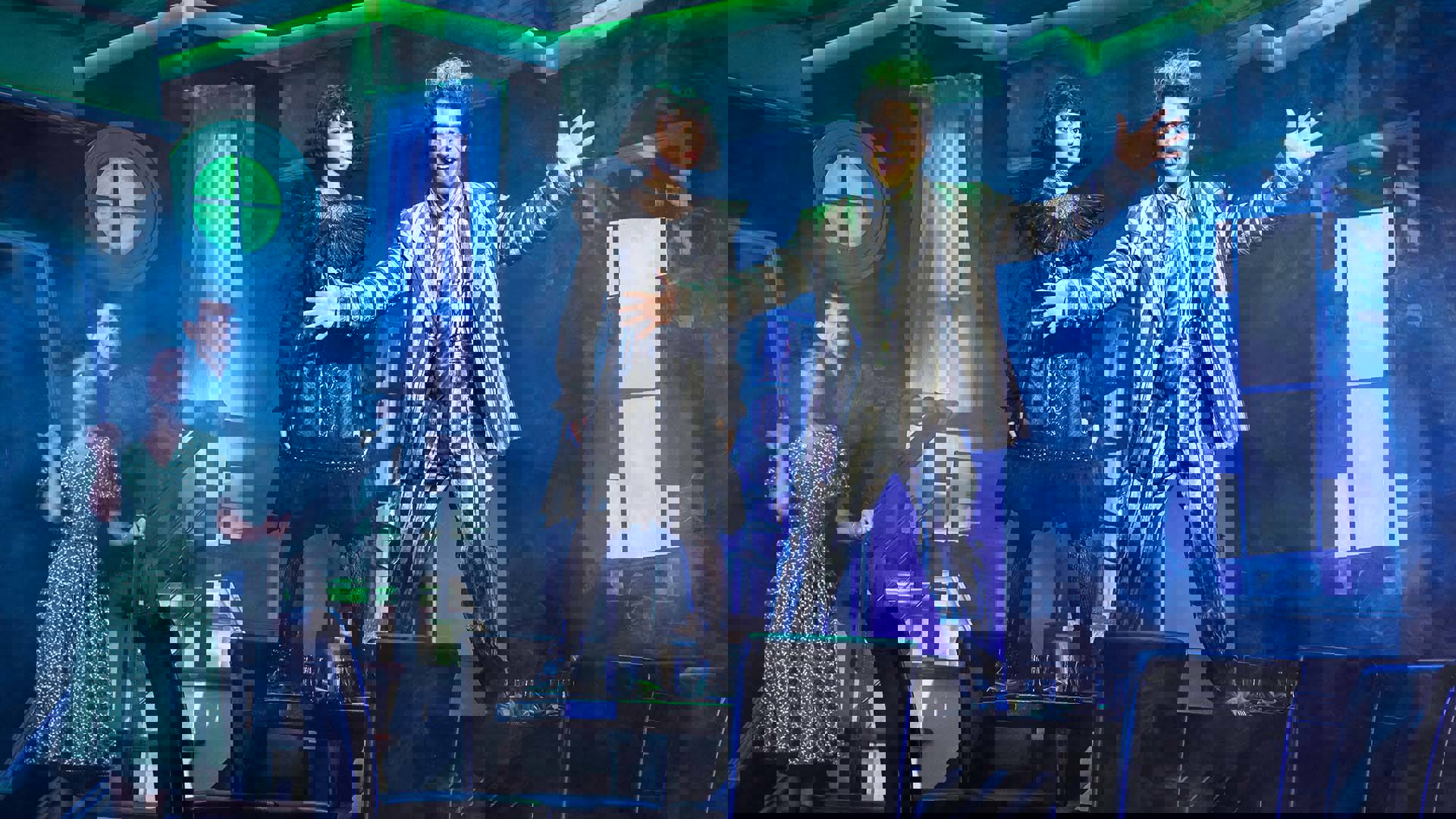 Beetlejuice and Lydia standing on a table with two people behind them. Beetlejuice is in a striped suit with his arms out wide, smiling.