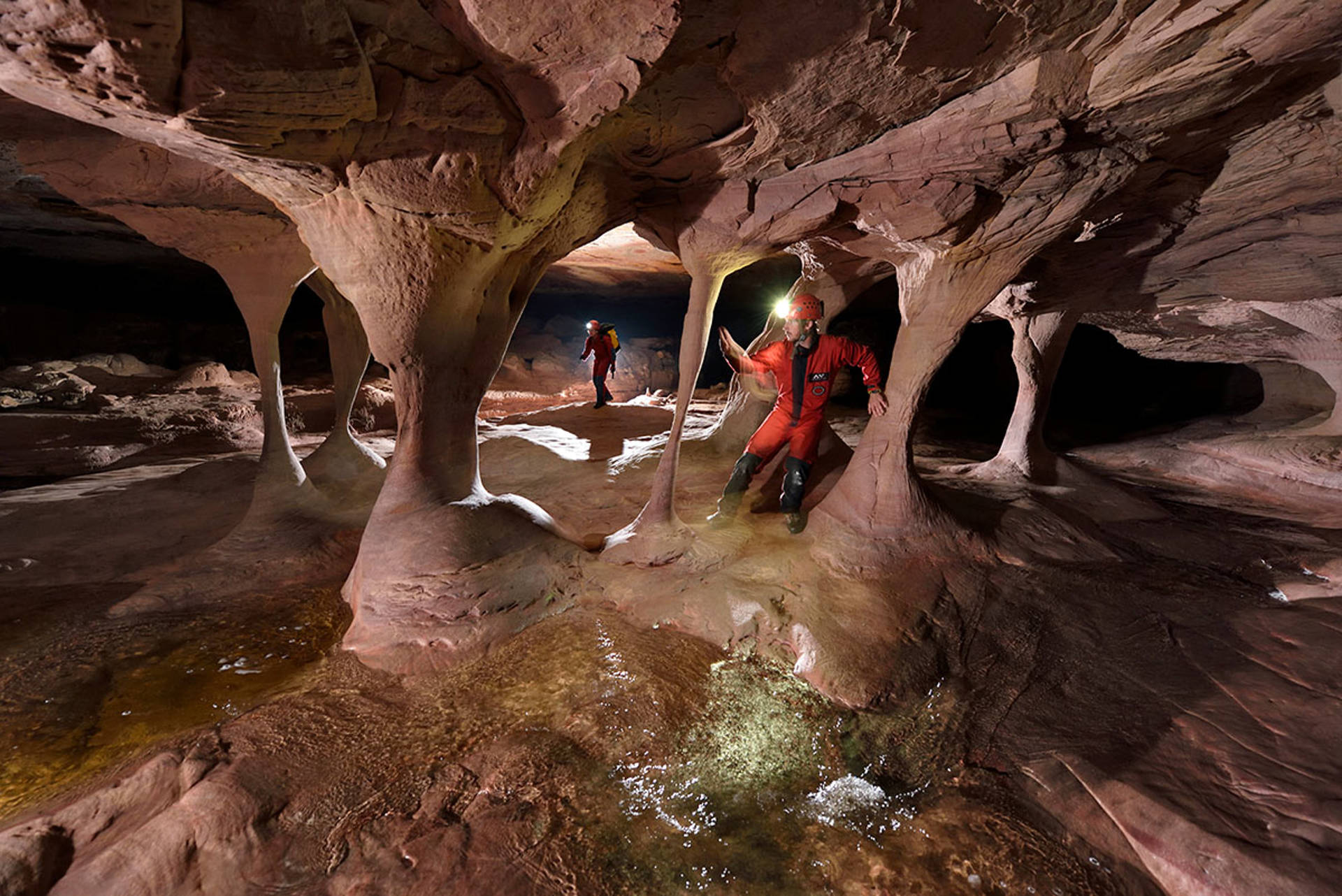 Two people in red outfits with headlamps explore a vast rust colored cave