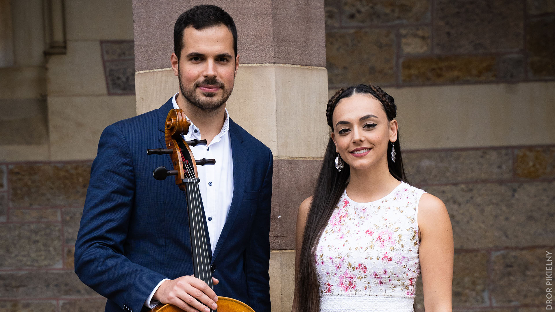 A man with light complexion, dark hair and a light beard in a blue suit coat propping up a cello. To his left is a young woman with light complexion, long dark hair, and a white dress.