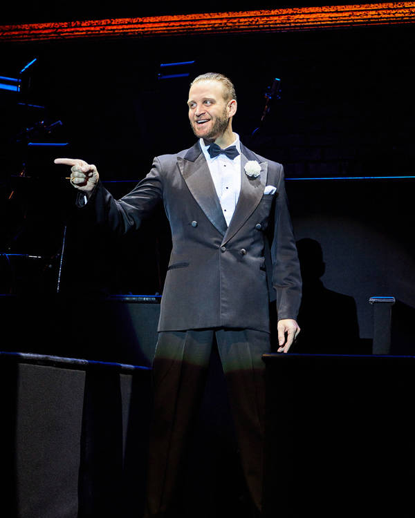 Jeff Brooks as Billy Flynn. A caucasian man stands on stage smiling wearing a classic tuxedo with one hand pointing out towards the audience.