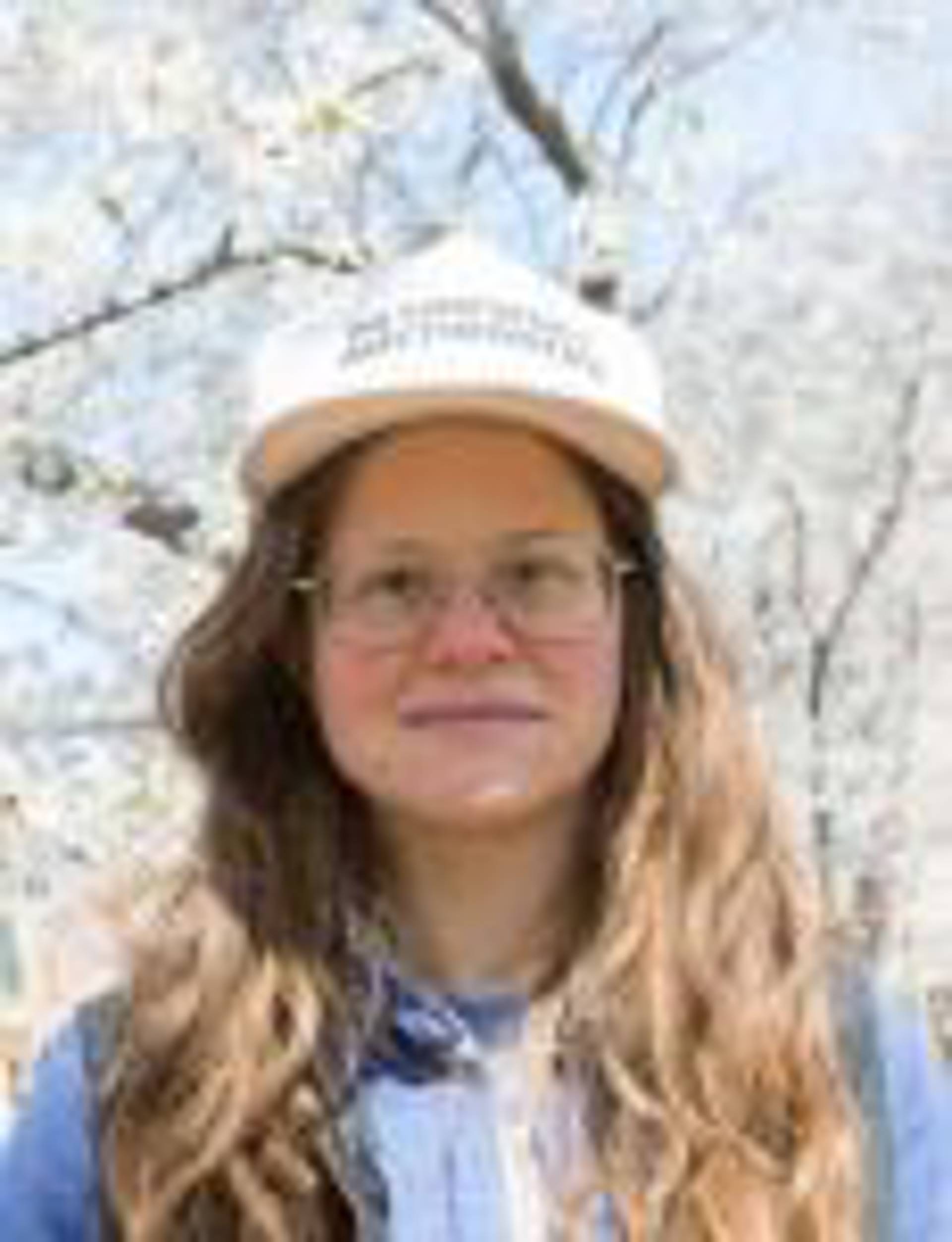 Headshot of a caucasian woman outside with long brown hair and a white hat on.