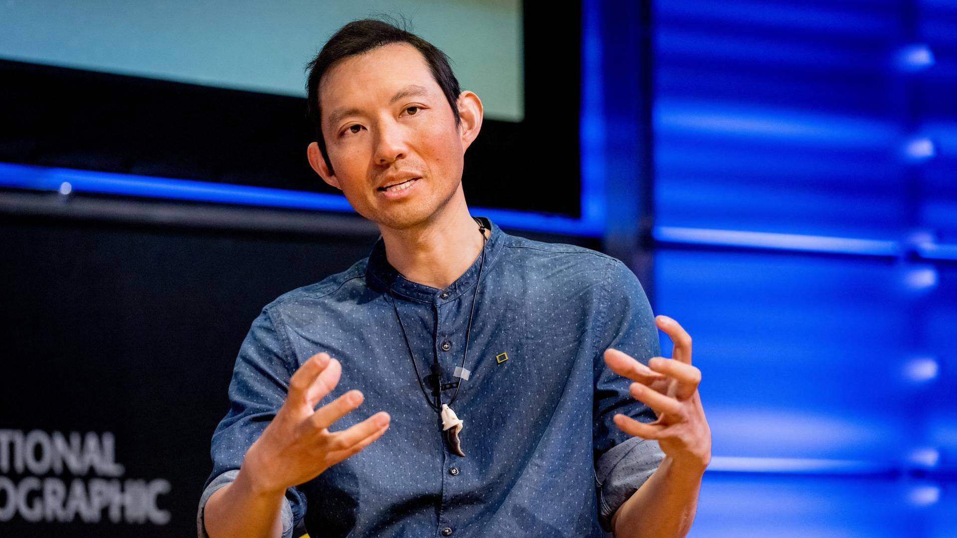 Photographer Kiliii Yuyan speaking in front of a black and blue background. He has dark hair and is wearing a blue shirt with white polka dots.