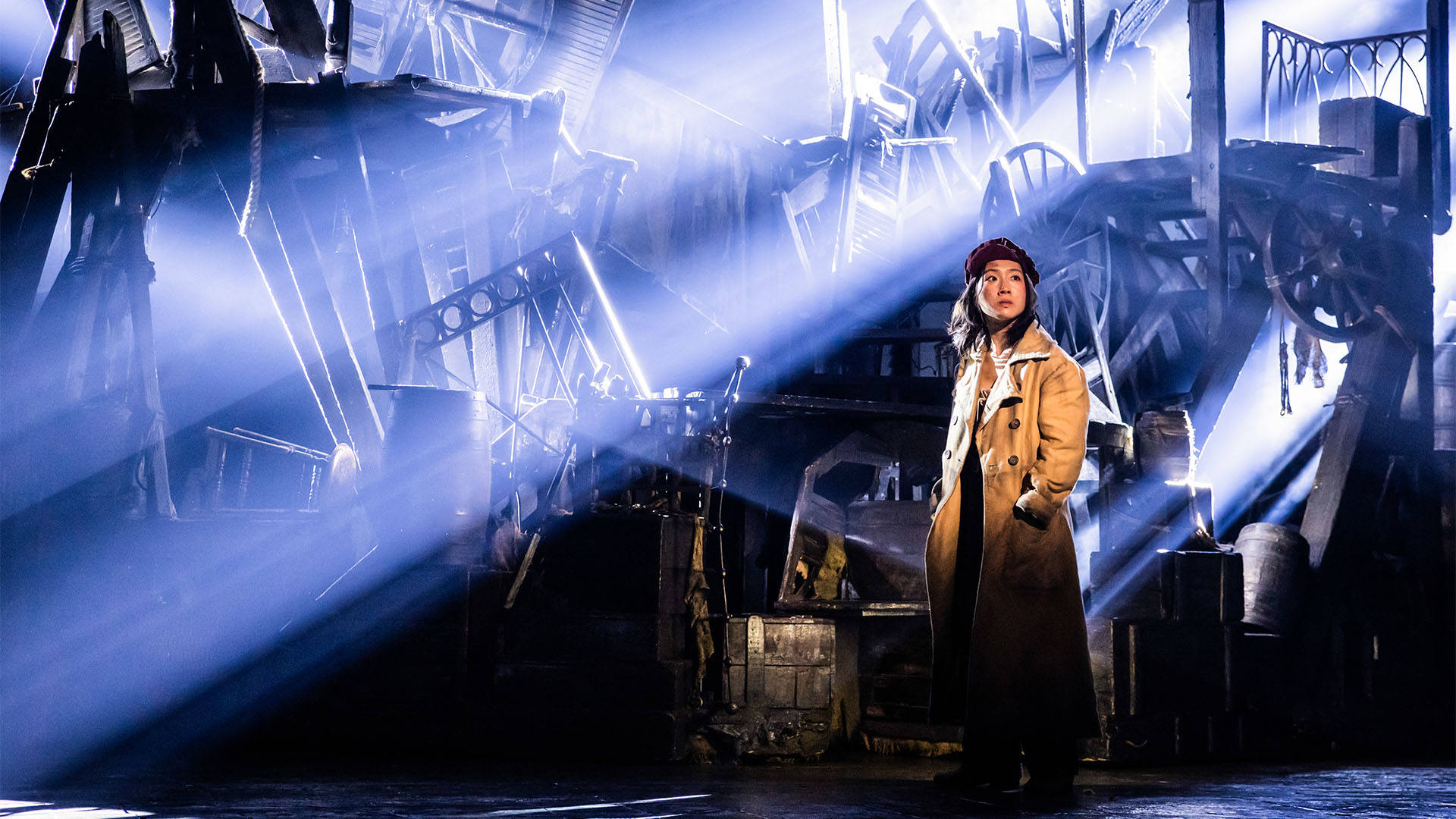 A woman in a long beige coat stands with a concerned look on her face.