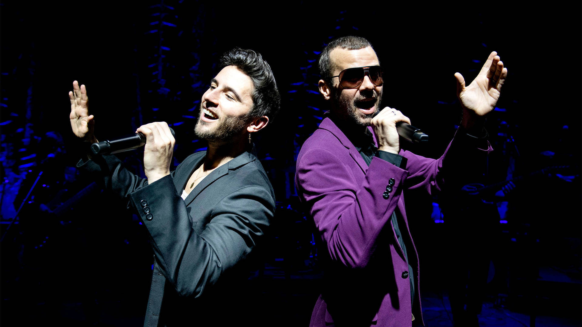 Two white men stand back to back singing into microphones. The man on the left has short brown hair and a dark jacket. The man on the right has buzzed dark hair, sunglasses and a purple jacket.