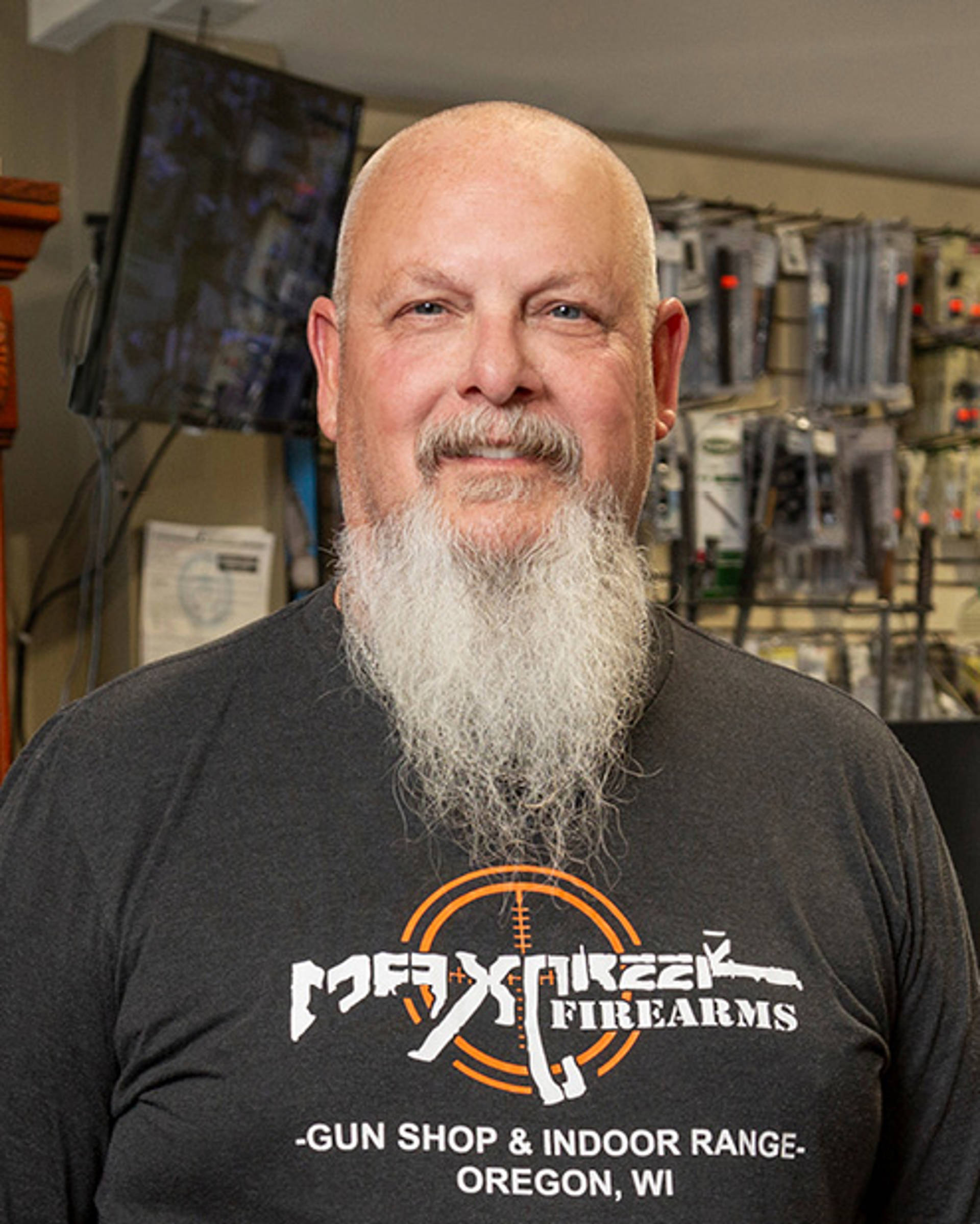 Headshot of a middle aged white man with a bald head and long grey bear. He is wearing a black Tshirt with words in the shape of an assault rifle, "MaxCreek Firearms"