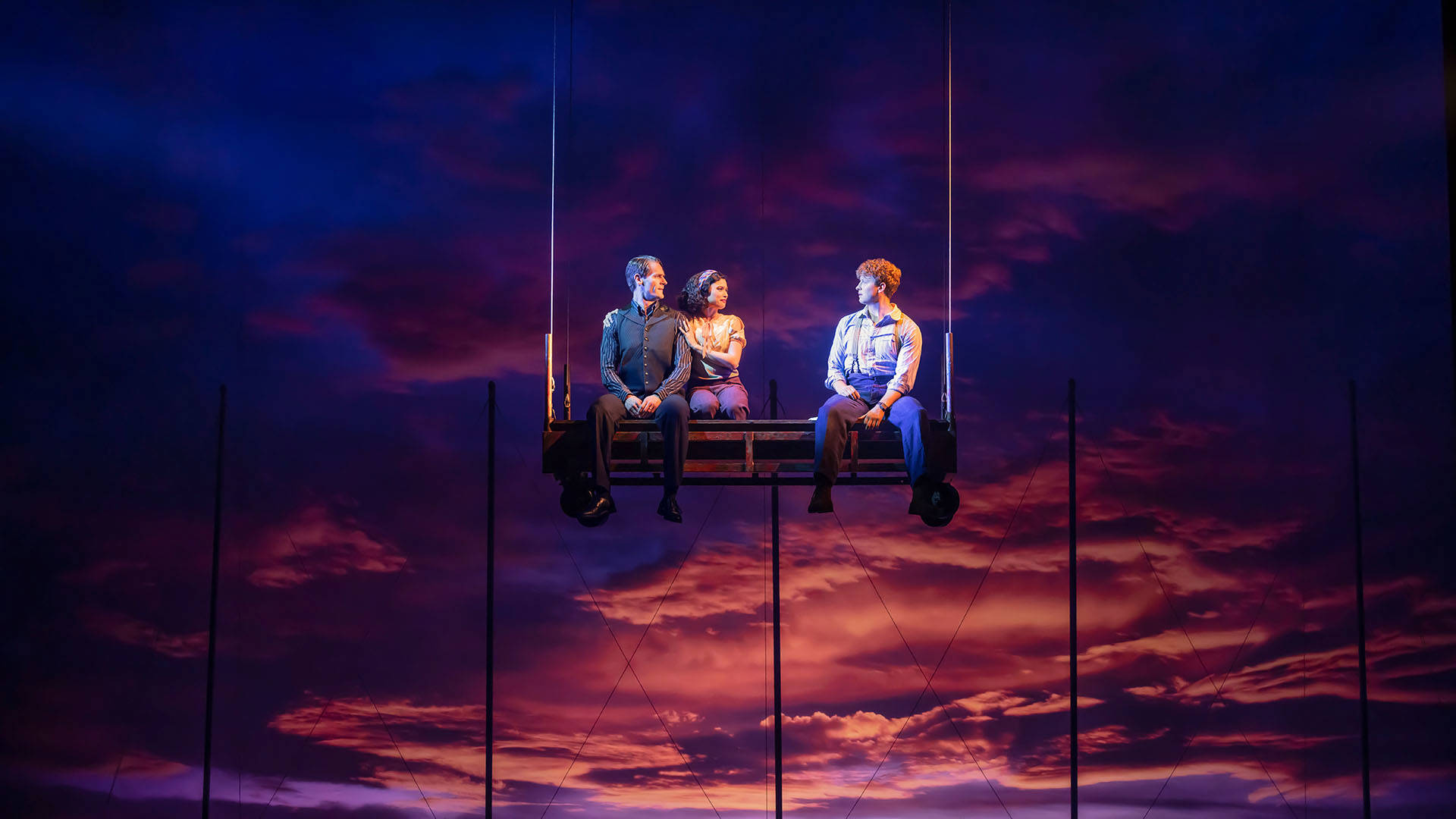Two men and a woman sit on a bench suspended in the air with a sunset backdrop.