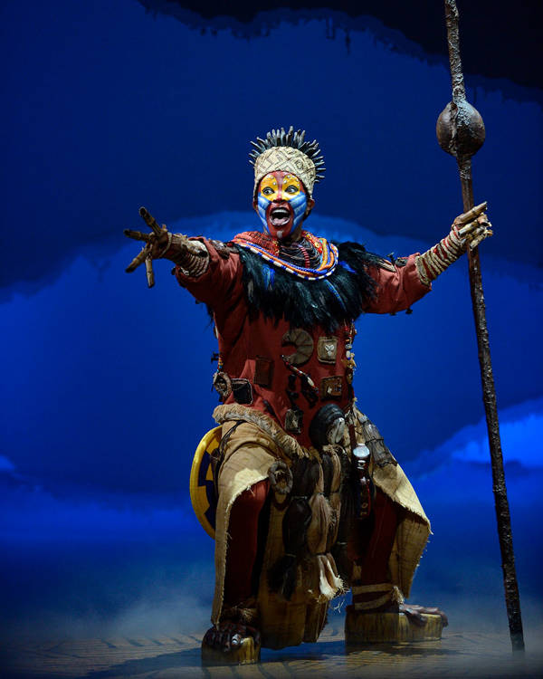 A woman dressed as Rafiki sings on stage.