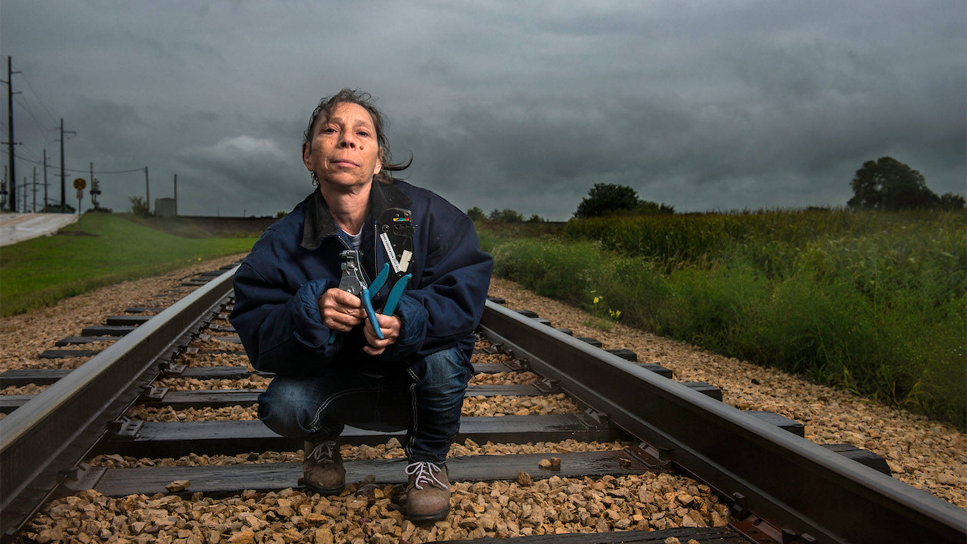 A portrait of a person with long brown hair on railroad tracks wearing a blue jacket, jeans and brown boots.
