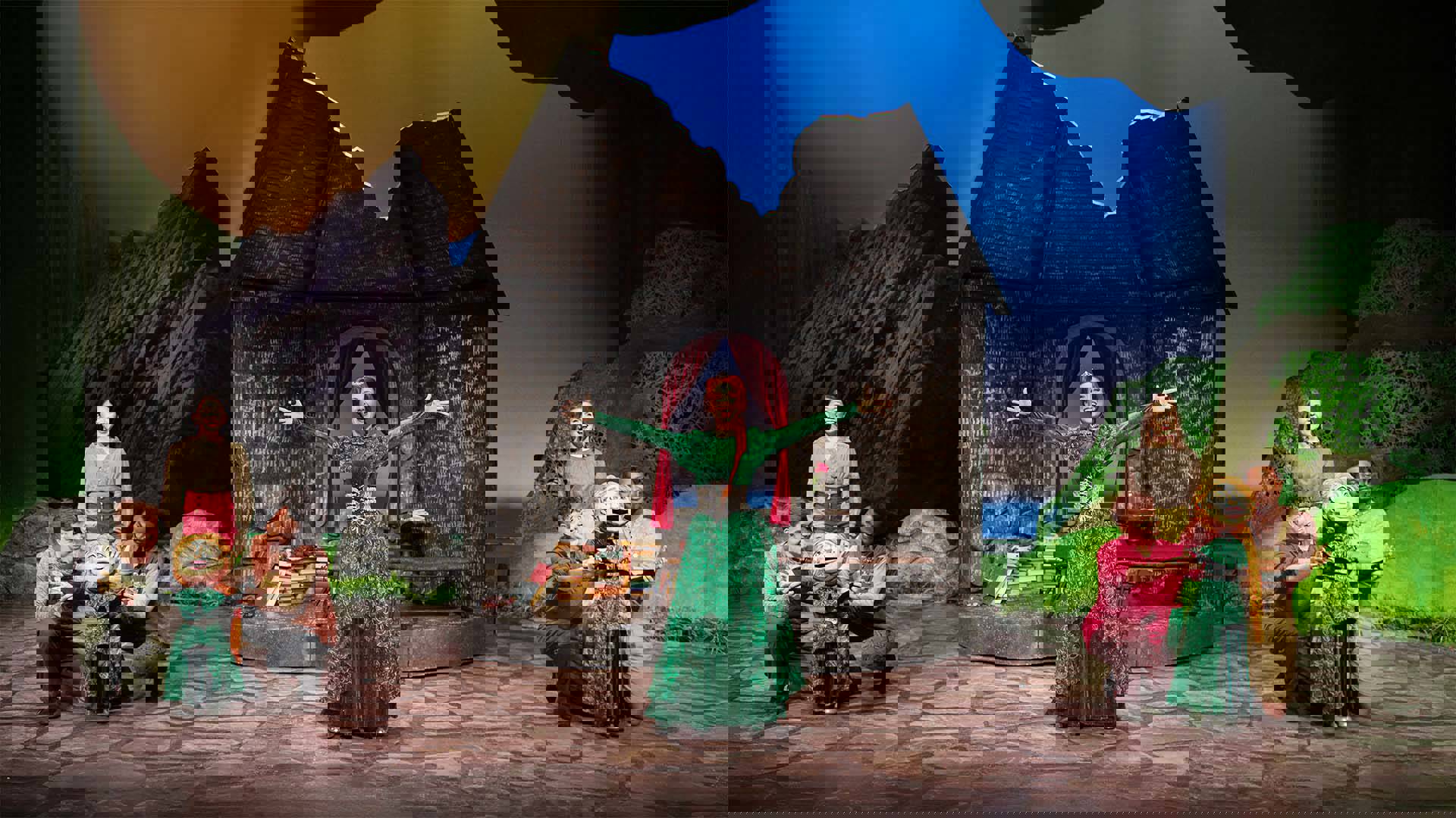 Princess Fiona, a white woman with a long red braid and tiara wearing a deep green gown standing outside her castle cell setpiece with a smile and arms raised open.