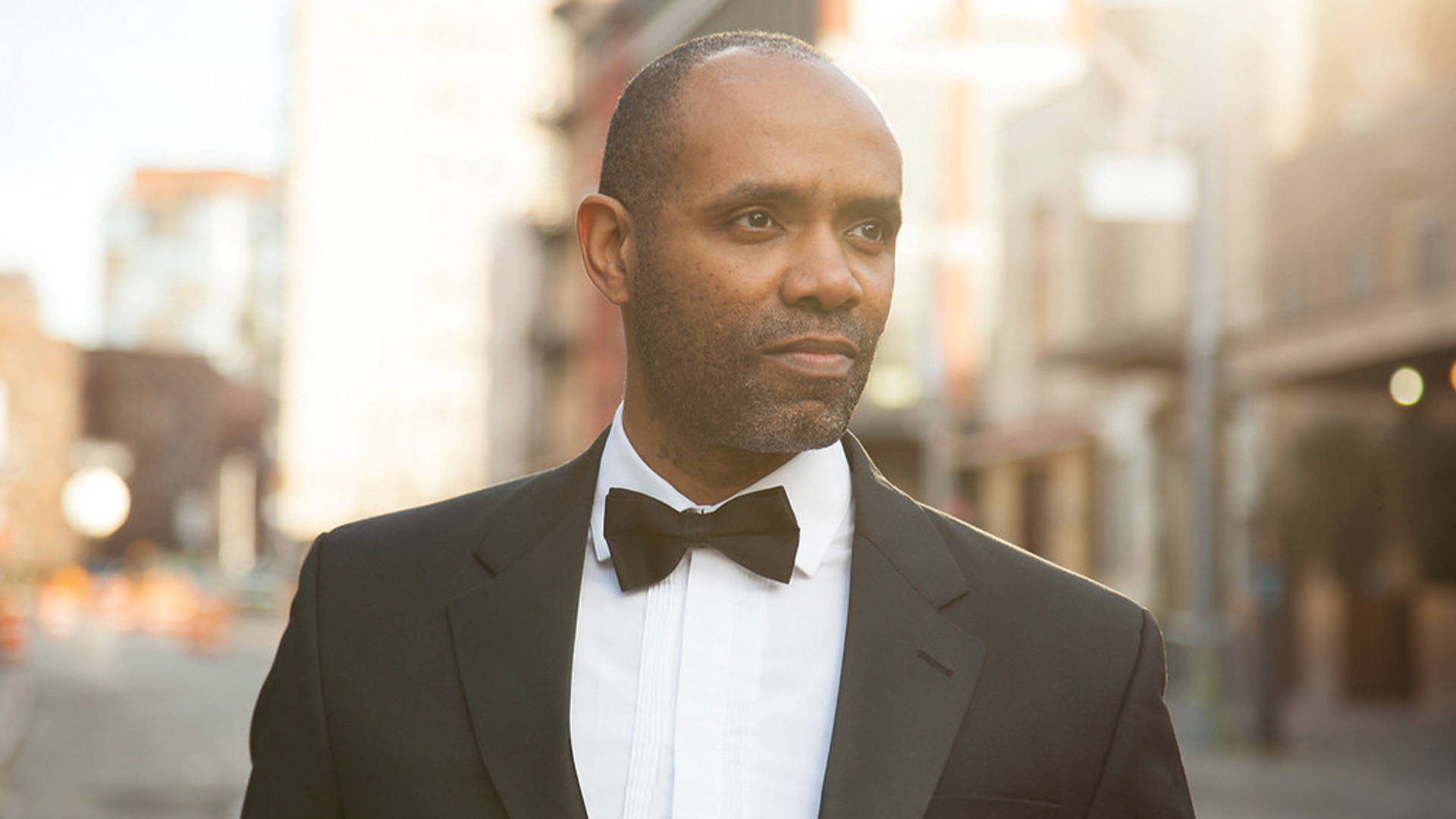 An African American man with short dark hair wearing a tuxedo. He has a stoic look on his face and there is a blurry city street behind him.