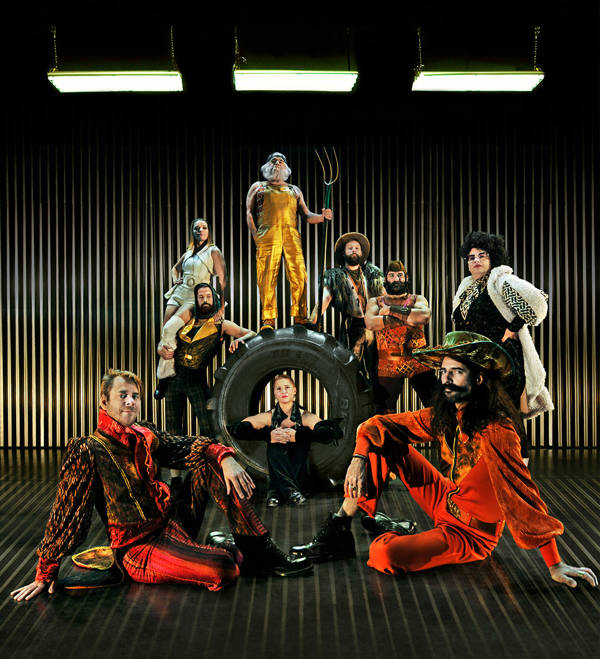 An incredible array of performers in funky futurist, somewhat steampunk outfits against a metalic background. One member is sitting in a giant tire and another is standing on top of it with a pitchfork
