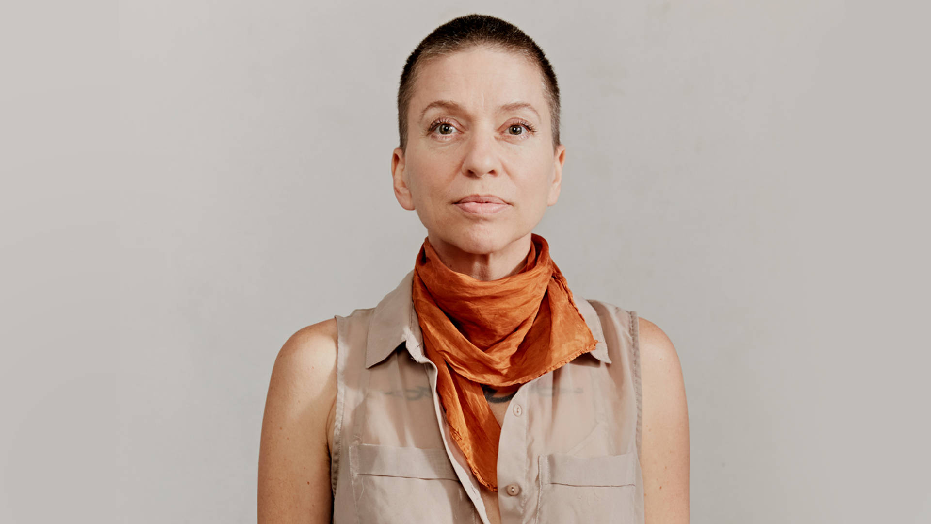 A middle-aged woman with light skin and buzzed, brown hair and brown eyes. She is wearing an orange scarf and a tan, sleeveless, button-down shirt.