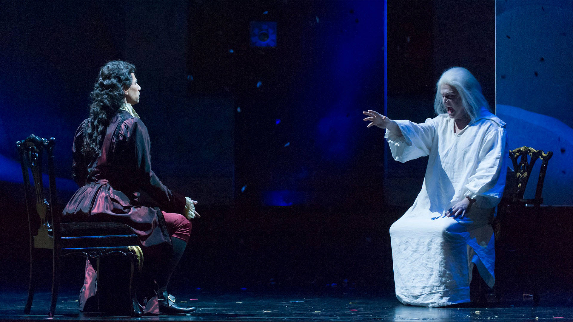 Performers in the opera "Don Giovanni" performing on a stage. A man in a dark and red outfit with long dark hair sits across from another man in all white with grey hair holding out his hand.
