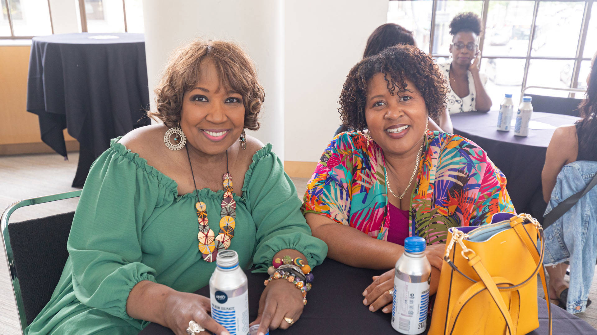 Two african american women seated at a table smiling. The woman on the left has brown eyes, brown wavy hair and a green dress. The woman on the right has shorter dark curly hair, brown eyes and a colorful top.
